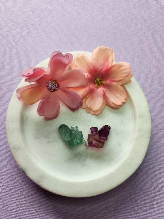 Close-up photo of two fluorite mini fairy carvings displayed in a white dish with two soft flower props on a lavender backdrop. The fairies show natural purple, green and blue fluorite banding with a polished glossy finish. Ideal for fluorite carvings, mini crystal fairies, whimsical crystal decor and collectible gemstone figures