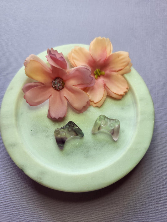 Close-up photo of two fluorite mini game controller carvings displayed in a white dish with two soft flower props on a lavender backdrop. Each controller shows natural purple, green and blue fluorite banding with a smooth polished finish. Perfect for fluorite carvings, gamer-themed crystal decor, mini crystal collectibles and unique gemstone gifts