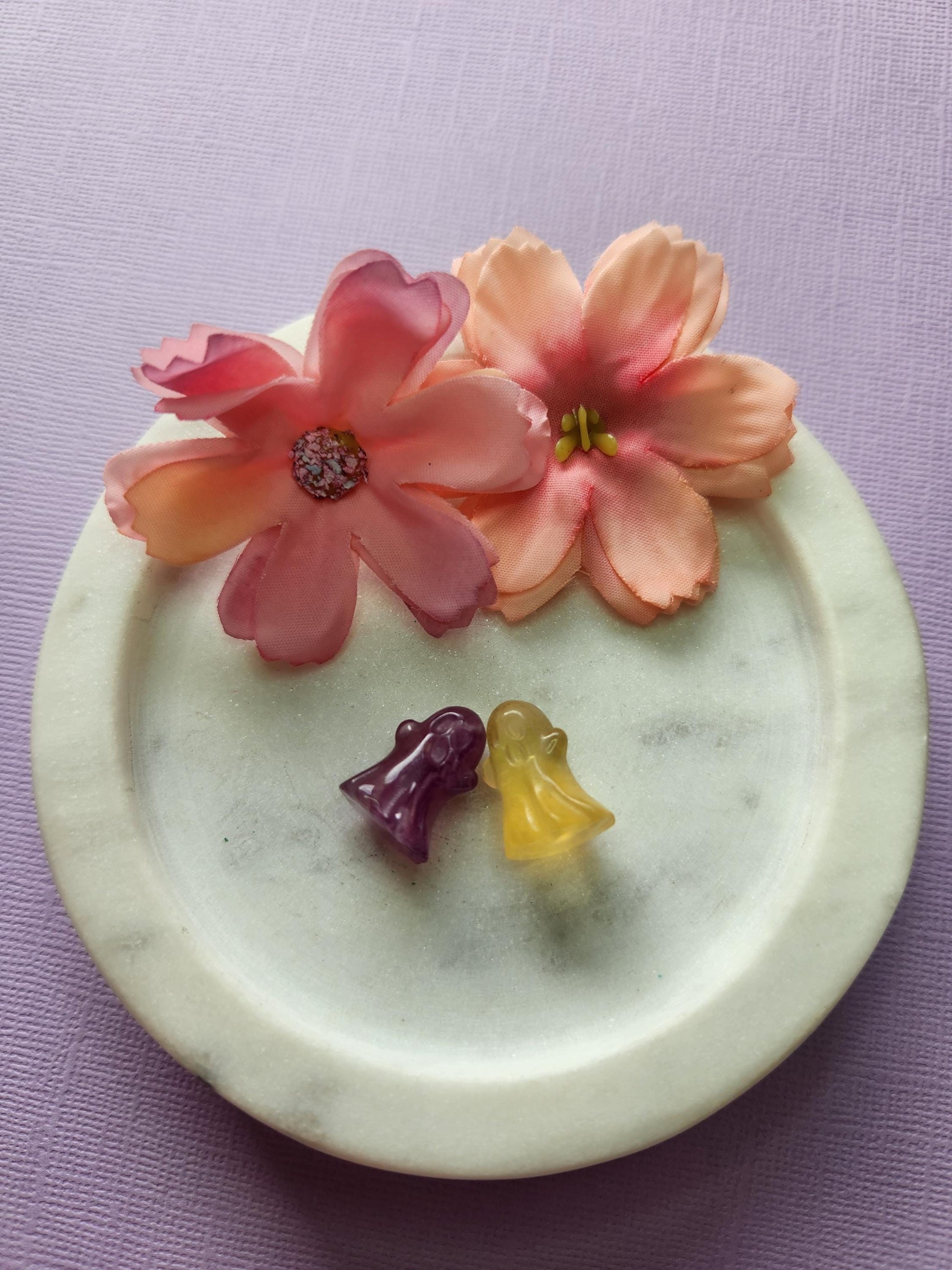 Close-up photo of two fluorite mini ghost carvings displayed in a white dish with two soft flower props on a lavender backdrop. The ghosts show natural purple, green and blue fluorite banding with a smooth polished finish. Ideal for fluorite carvings, mini crystal ghosts, cute crystal decor and collectible gemstone figures