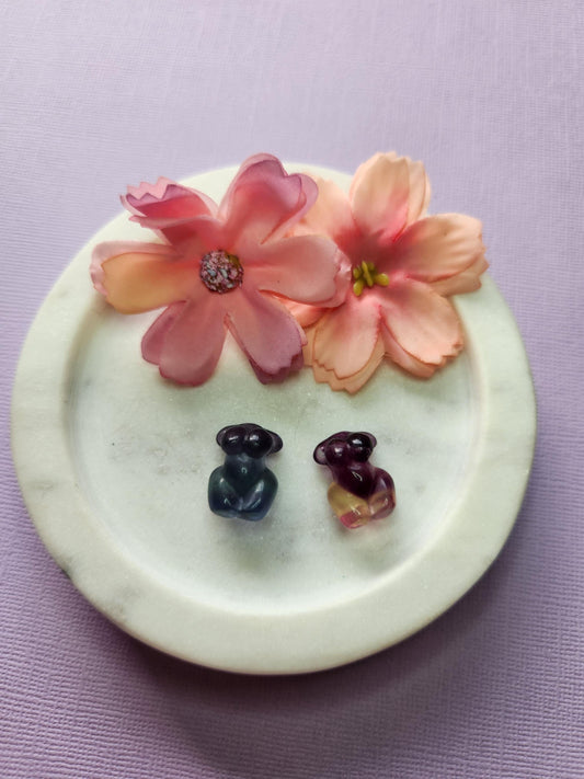 Close-up photo of two fluorite mini goddess body carvings displayed in a white dish with two soft flower props on a lavender backdrop. The carvings show natural purple, green and blue fluorite banding with a smooth polished finish. Ideal for fluorite carvings, goddess body crystals, mini crystal decor and collectible gemstone figures