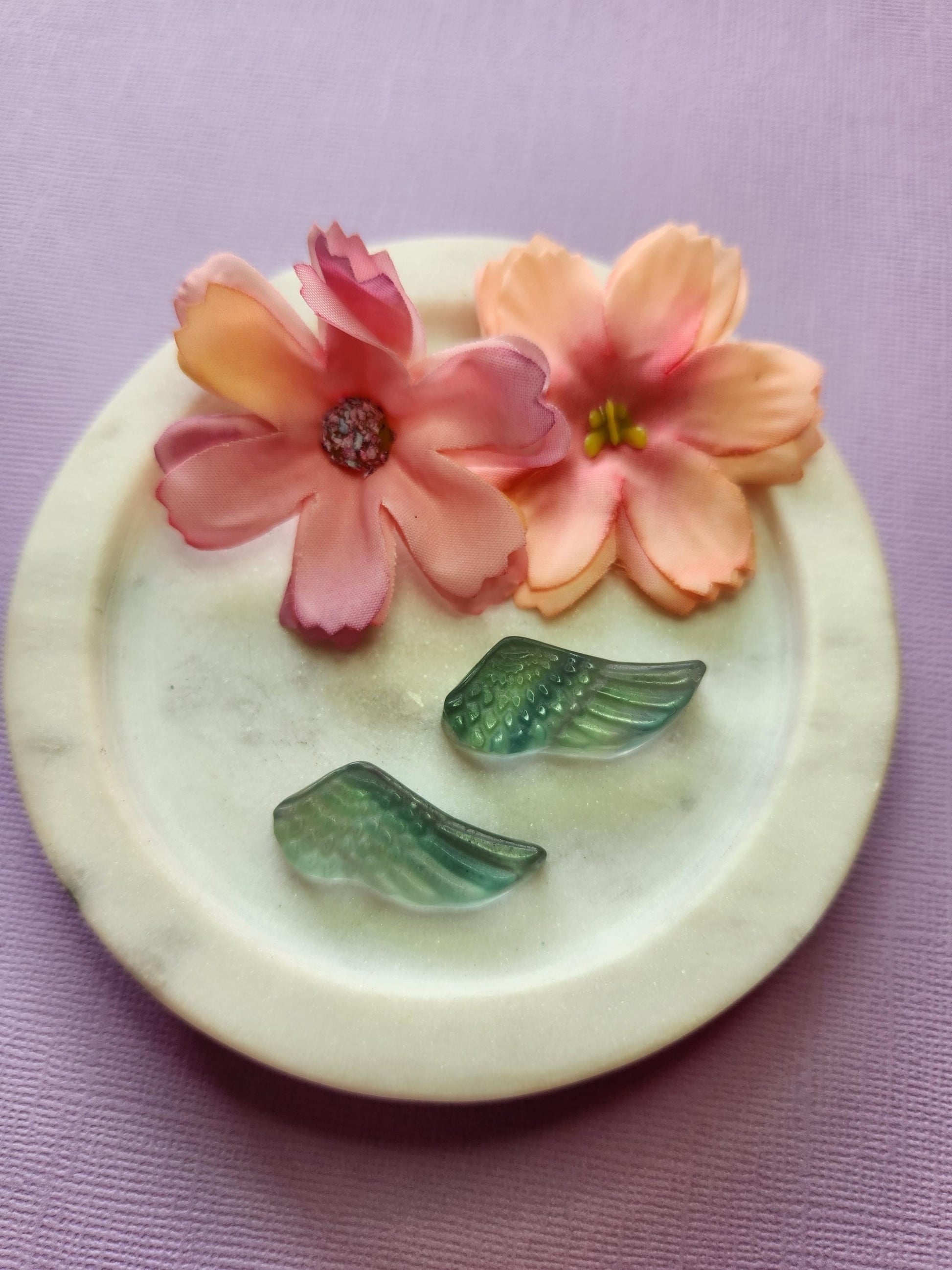 Close-up photo of two fluorite mini wing carvings displayed in a white dish with two soft flower props on a lavender backdrop. The wings show natural purple, green and blue fluorite banding with a smooth polished finish. Ideal for fluorite carvings, angel wing crystals, mini crystal decor and collectible gemstone accents