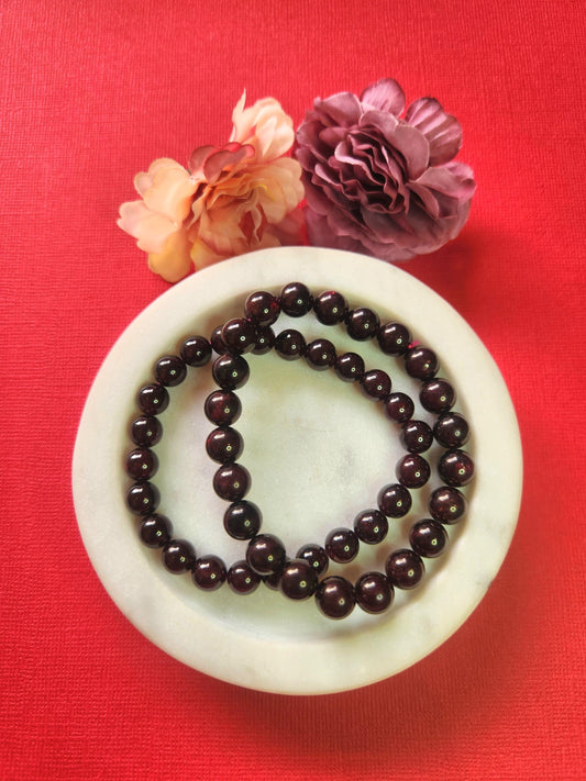 Garnet 8mm beaded crystal bracelet displayed in a white dish with two flower props on a rich red background, deep red gemstone bracelet for grounding, passion and strength, healing crystal jewelry photo for energy and vitality.
