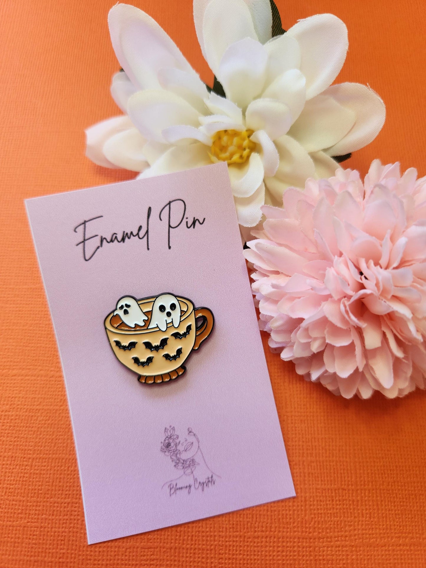 Orange enamel coffee cup pin with black bat silhouettes on the cup and two white ghosts rising from the top of the coffee. Styled with two flower props on a bright orange background. Ghost coffee pin, spooky cute enamel pin, Halloween coffee cup accessory, bat and ghost pin, cozy witchy aesthetic lapel pin.