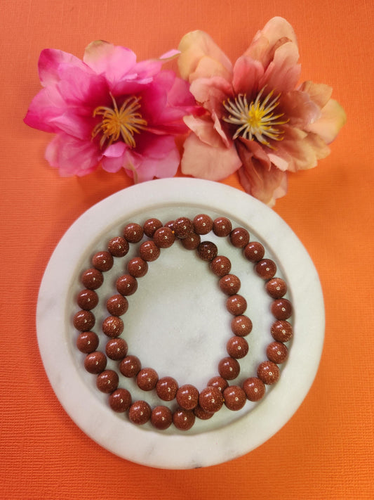 Two 8mm Gold Sandstone beaded bracelets in white dish with two flower props on orange background, natural gold sandstone gemstone beads, crystal jewellery, handmade beaded bracelet for grounding, protection and energy healing, gemstone accessory product photo