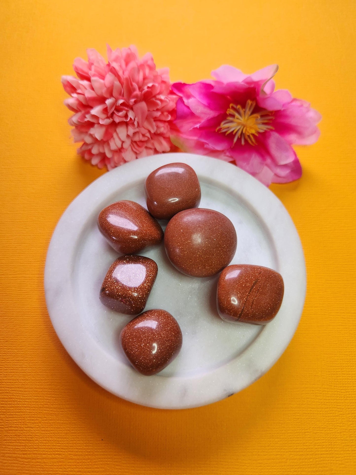 Gold sandstone tumbles in a white dish with two flower props on a yellow background, sparkling gold sand stone crystal tumbles for confidence motivation and energy, glittery man-made goldstone healing stones product photo