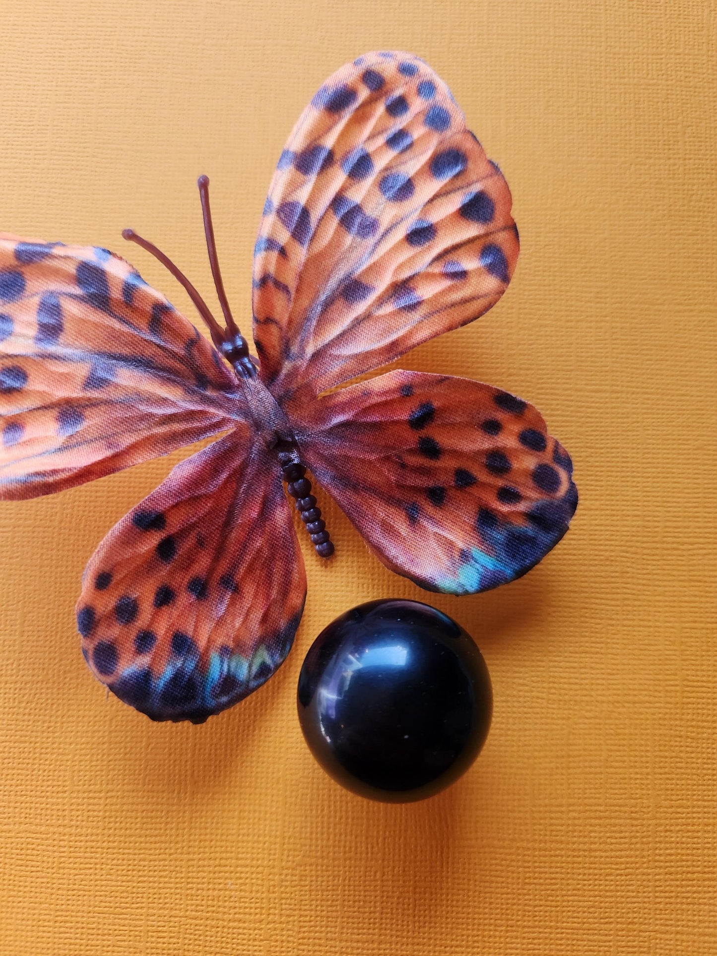 Gold Sheen Obsidian Sphere displayed on an orange-gold background with an orange butterfly nearby. This polished crystal ball features a shimmering gold sheen, perfect for energy work, protection, grounding, and shadow work practices. Ideal for witchy decor, altar setups, and crystal collectors. The butterfly adds a touch of transformation symbolism, complementing the obsidian's deep metaphysical energy.