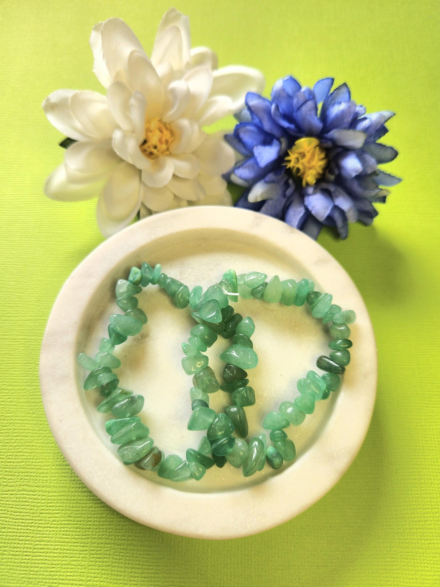 Two Green Aventurine crystal chip bracelets displayed in a white dish with delicate flower props on a bright green background. Handmade gemstone bracelets for luck, prosperity, and heart chakra healing, perfect crystal jewelry gift idea.