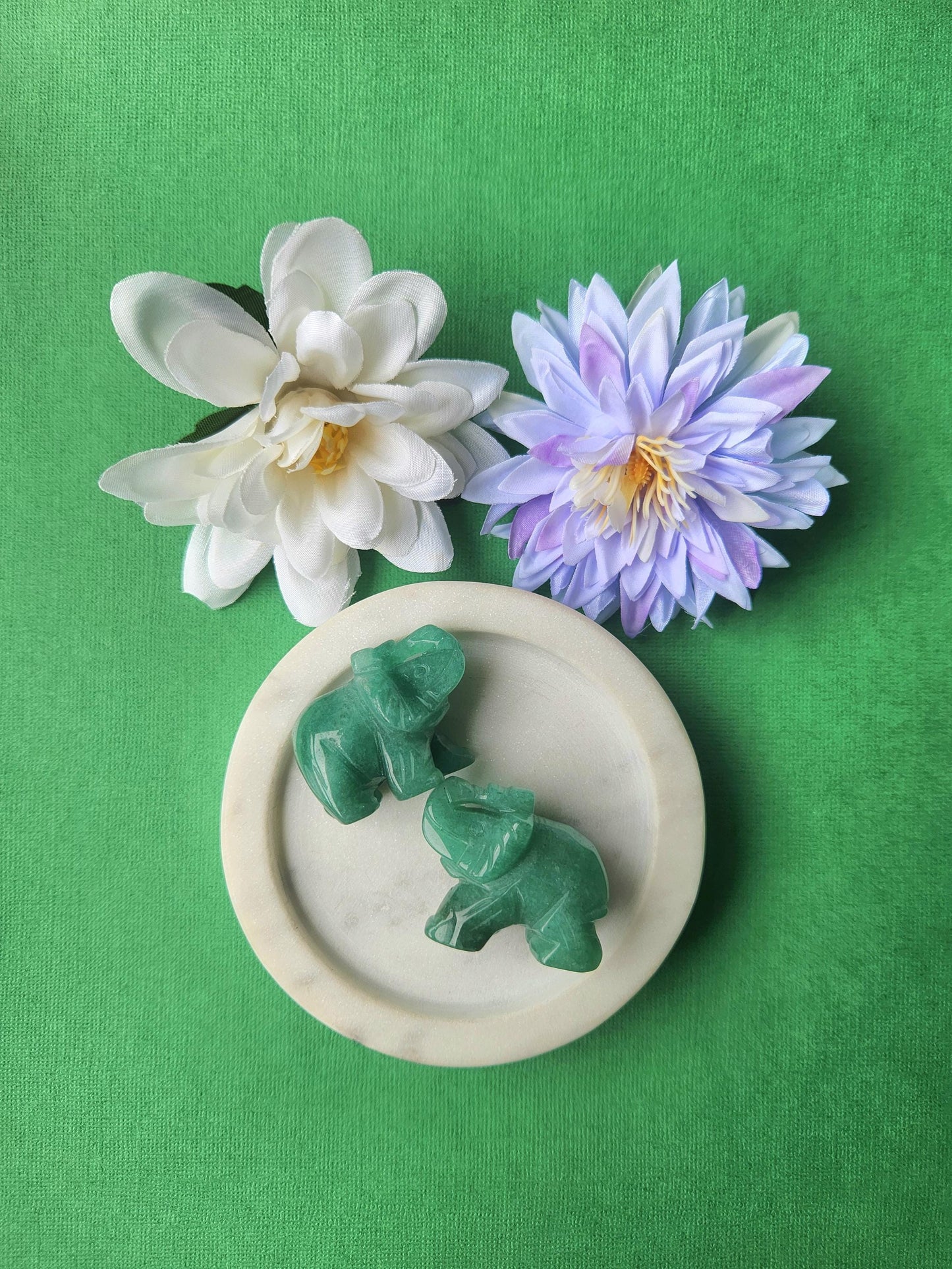 Green aventurine elephant crystal carvings displayed together in a white dish with floral props on a green background, polished gemstone elephant figurines for luck, abundance, crystal decor, and gifting