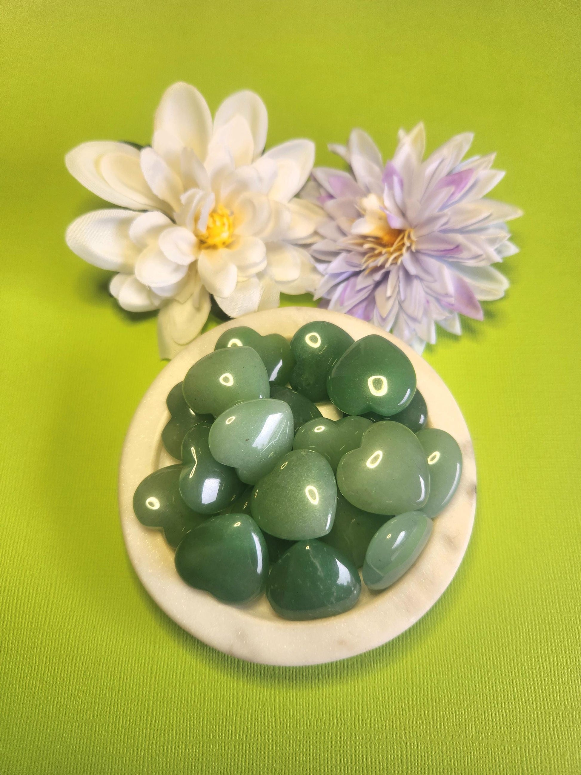 Green aventurine crystal heart displayed in a white dish on a green background with soft flower props featuring polished heart stone for luck abundance love healing crystal decor and gift