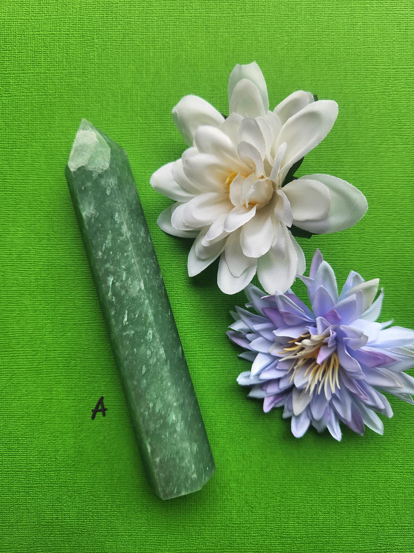 Green aventurine crystal tower A standing upright on a green background with delicate flower props highlighting polished green aventurine point for luck abundance and crystal healing decor
