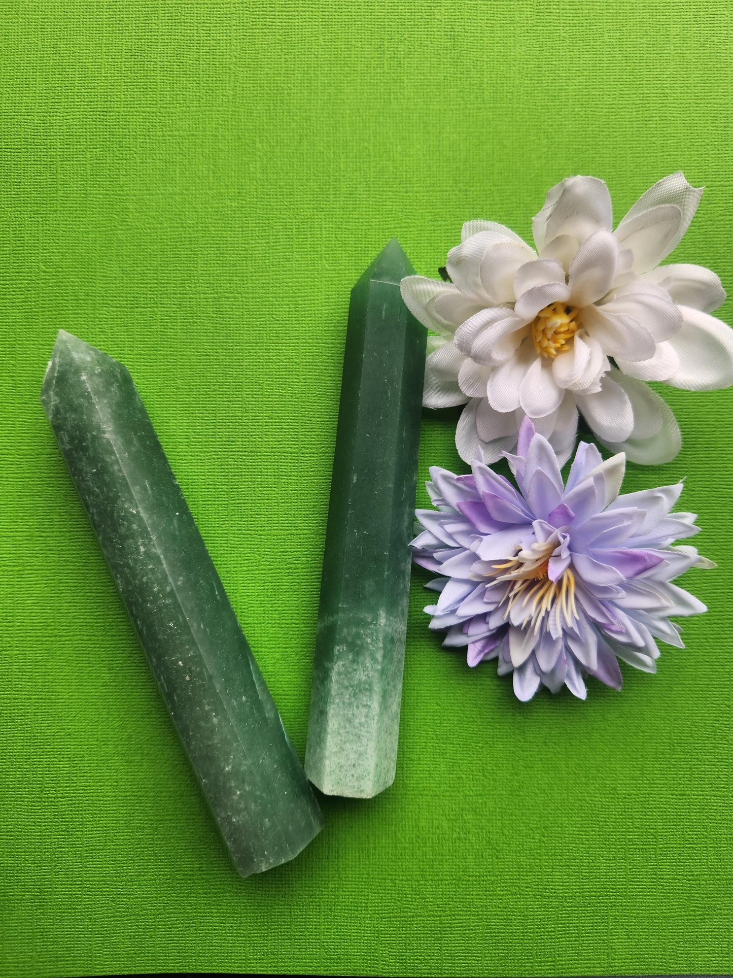 Green aventurine crystal towers pair displayed upright on a green background with soft flower props featuring polished green stone points for crystal decor abundance and healing crystals