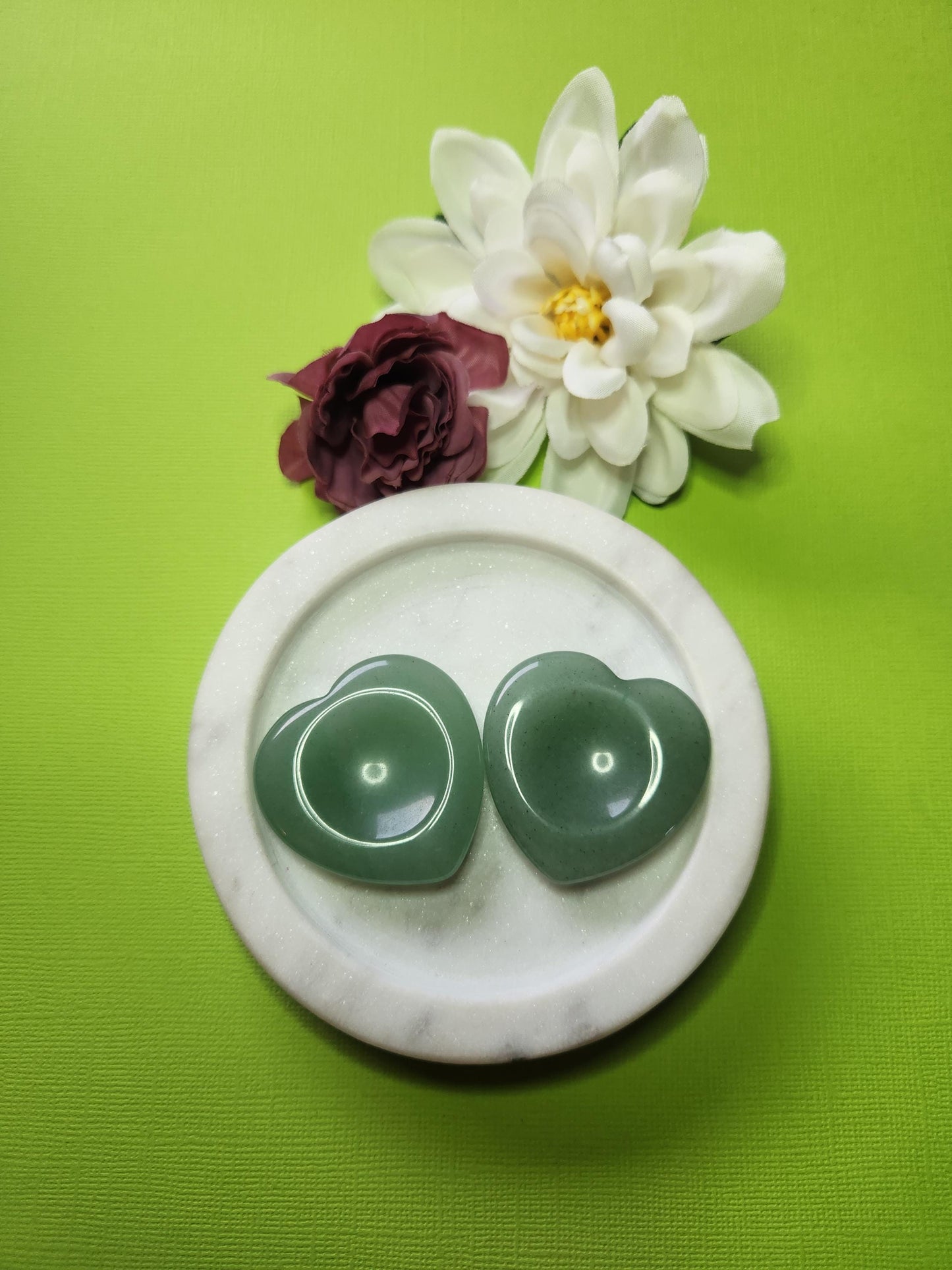 Two Green Aventurine heart-shaped worry stones displayed in a white dish with two flower props on a green background, polished pocket crystals for luck, prosperity, and stress relief.