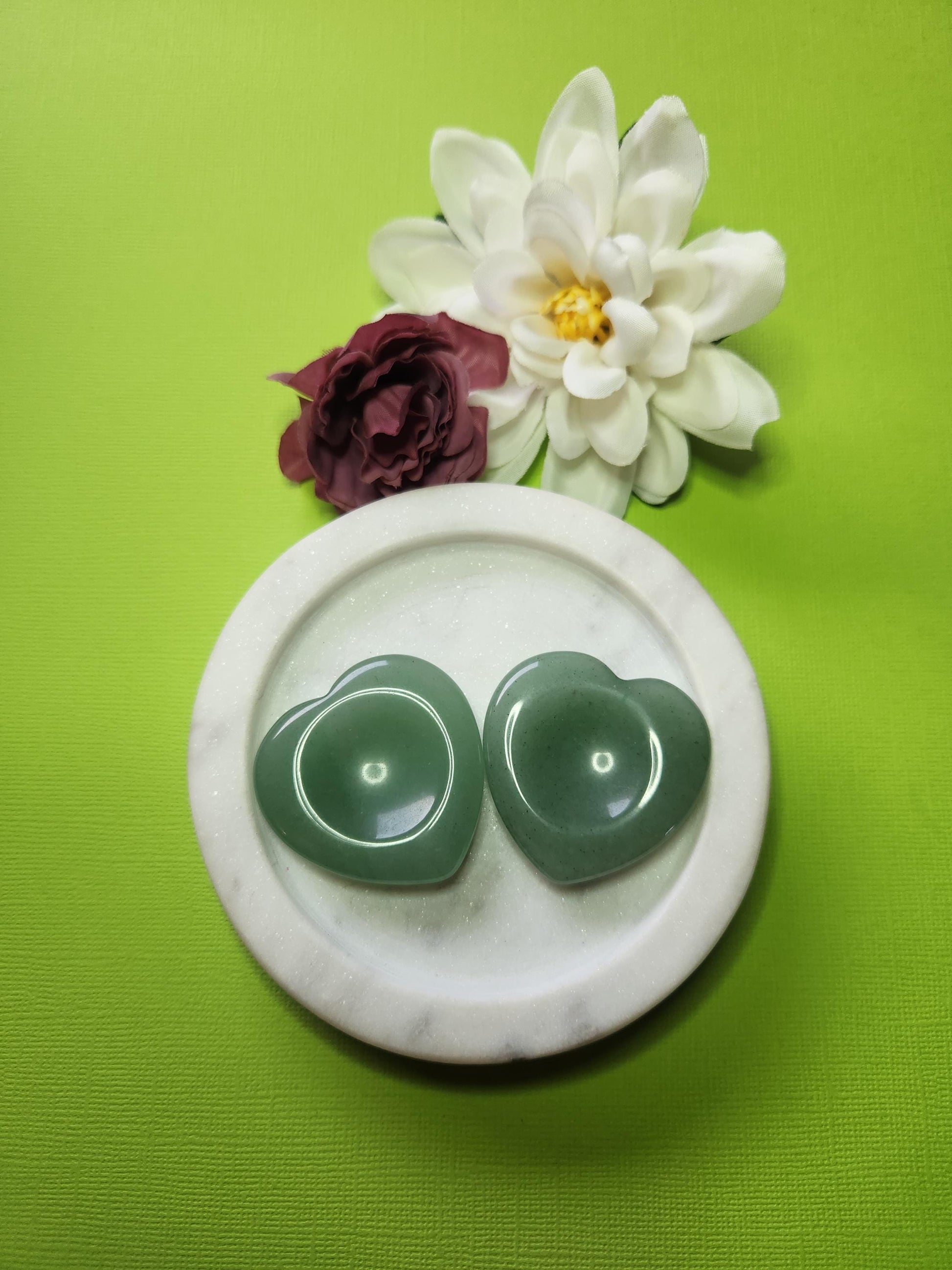 Two Green Aventurine heart-shaped worry stones displayed in a white dish with two flower props on a green background, polished pocket crystals for luck, prosperity, and stress relief.