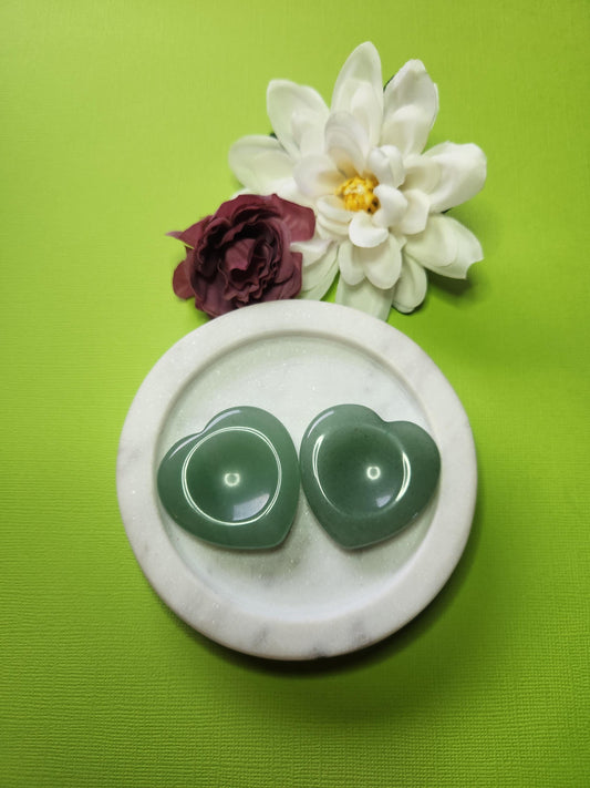Two Green Aventurine heart-shaped worry stones displayed in a white dish with two flower props on a green background, polished pocket crystals for luck, prosperity, and stress relief.