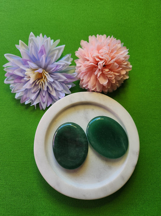 Two polished Green Aventurine worry stones displayed on a white dish with two flower props, set against a green backdrop. Calming crystal thumb stones for stress relief, heart chakra healing, and emotional balance.