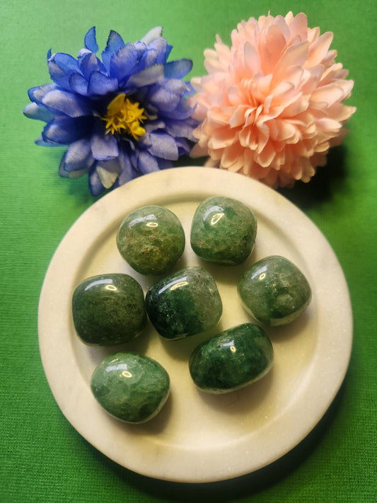 Green strawberry quartz tumble stones displayed in a white dish with two flower props, set against a green background. Polished natural quartz pieces known for heart-centered energy, emotional healing, and positivity — perfect for crystal collections, meditation, and spiritual décor.