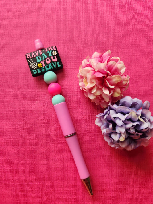 Have The Day You Deserve silicone beaded pen with black focal bead featuring skeleton hand peace sign and pink/blue lettering, pink and blue silicone beads on a pink pen, styled flat lay with two flower props on a pink background, aesthetic stationery photo for handmade pen shop, cute spooky pen gift, pastel gothic office supplies