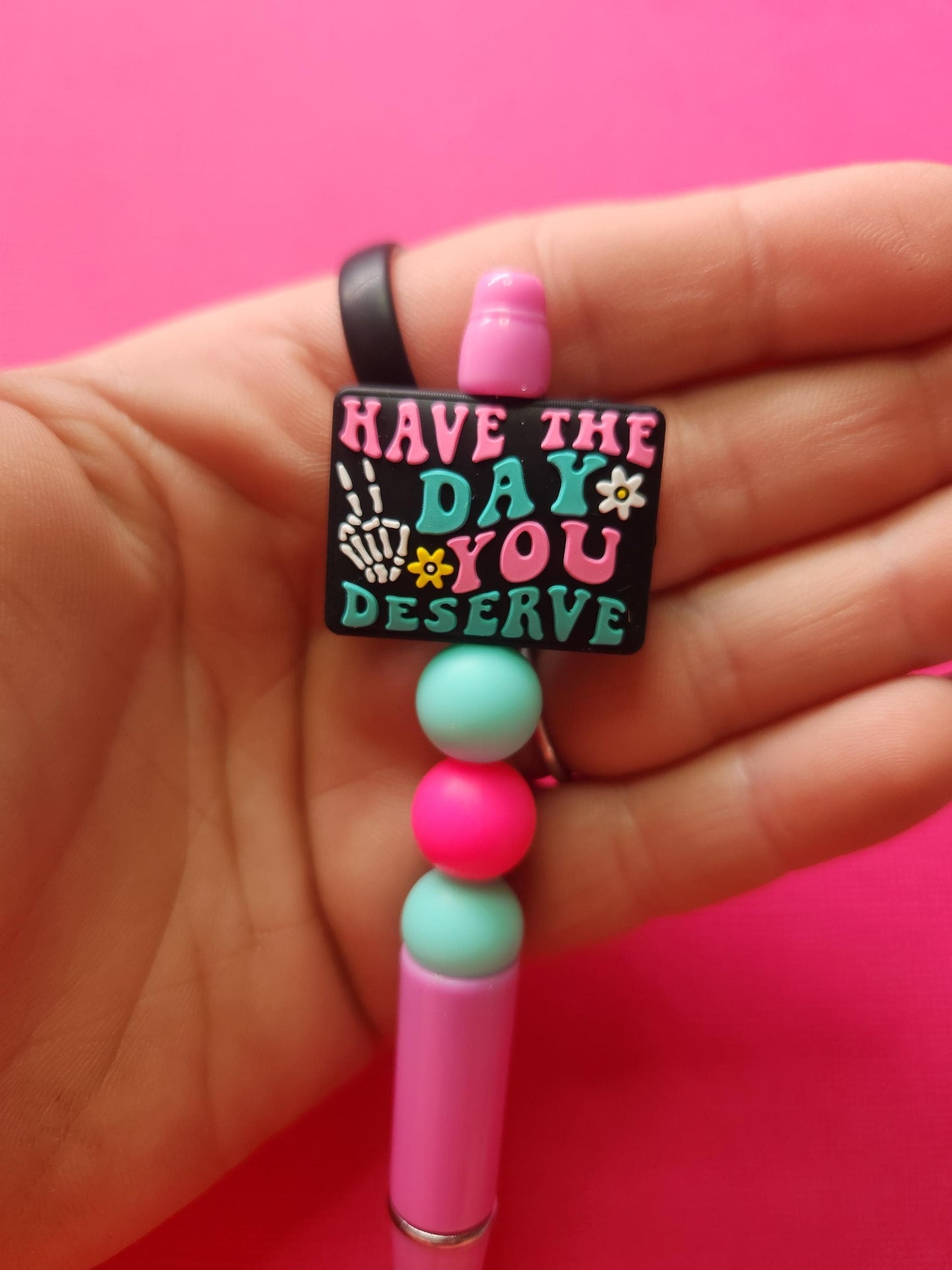 Close-up of Have The Day You Deserve silicone beaded pen held in hand showing black focal bead with skeleton peace sign and pink/blue text, pink and blue silicone accent beads on pink pen, detailed product shot on pink background for SEO, handmade spooky cute pen, pastel goth stationery