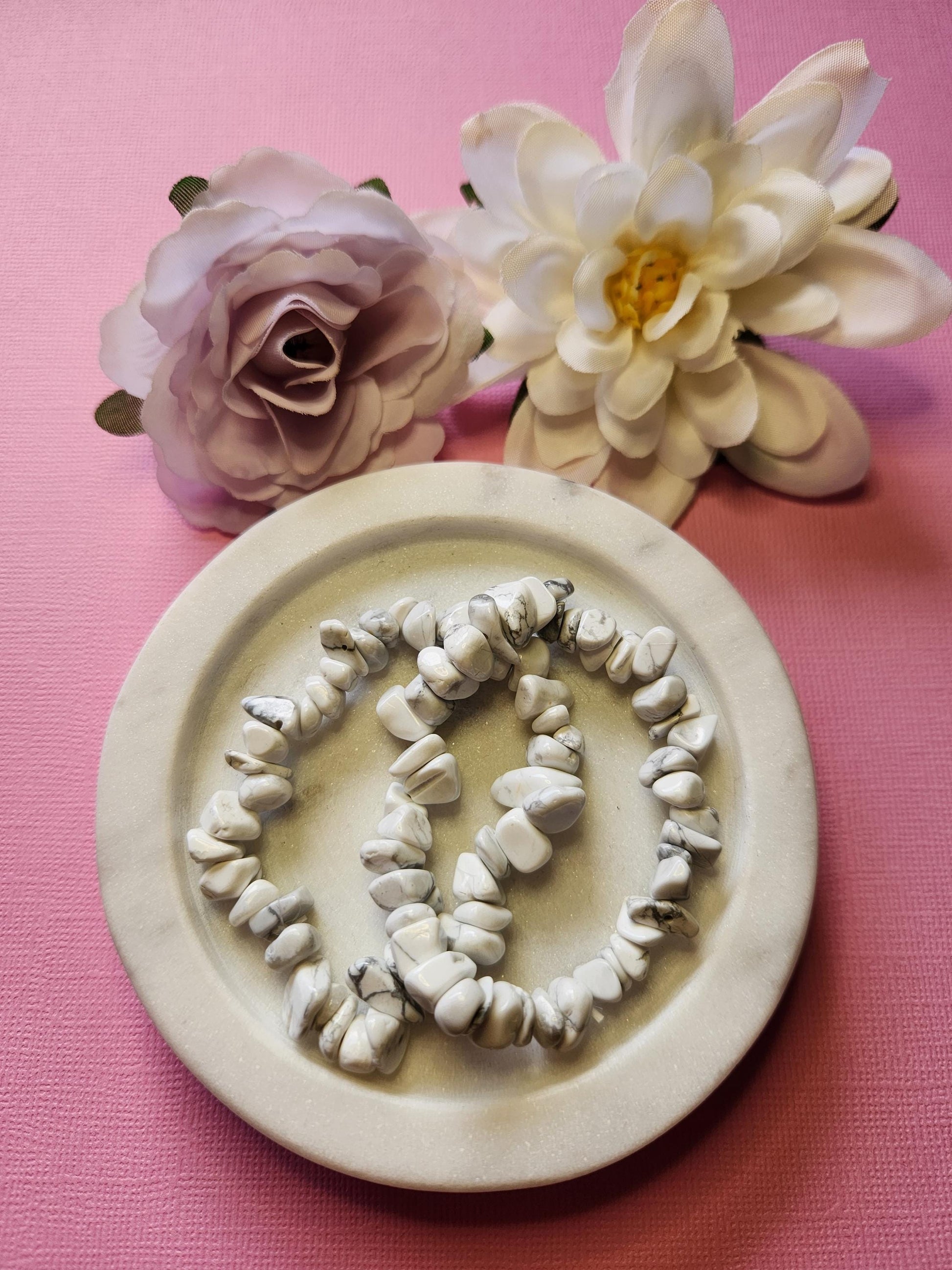 Two Howlite chip bracelets displayed in a white dish with two pink and white flower props on a light pink background. Stretch gemstone bracelets for mindfulness, meditation, stress relief, emotional balance, crown chakra healing, spiritual energy, and crystal jewelry gift for her