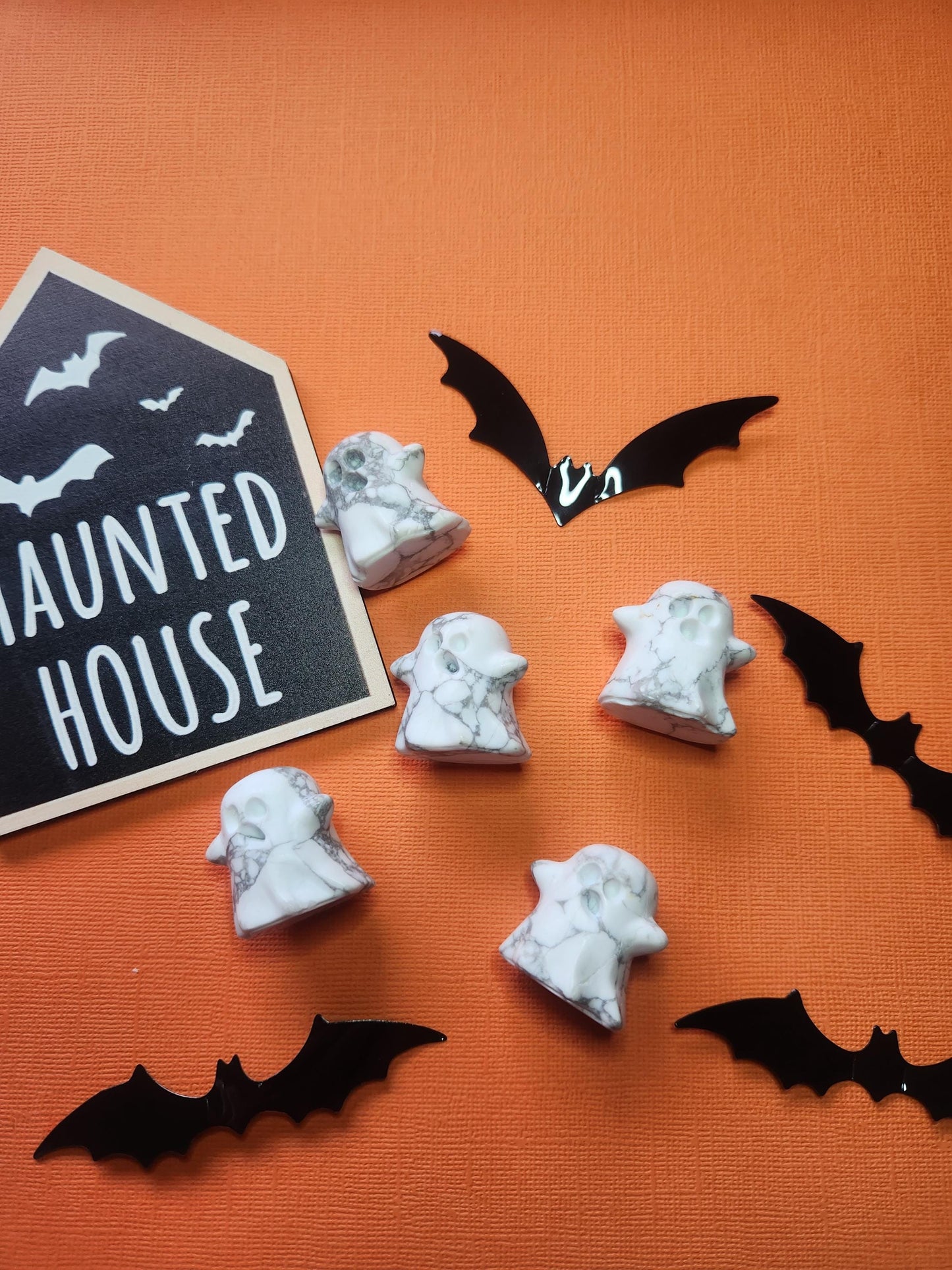 Flat lay photo featuring a white howlite ghost crystal surrounded by black bat cut-outs and a "Haunted House" sign, arranged on an orange backdrop. A Halloween-inspired scene with spooky, witchy vibes and contrasting colours.