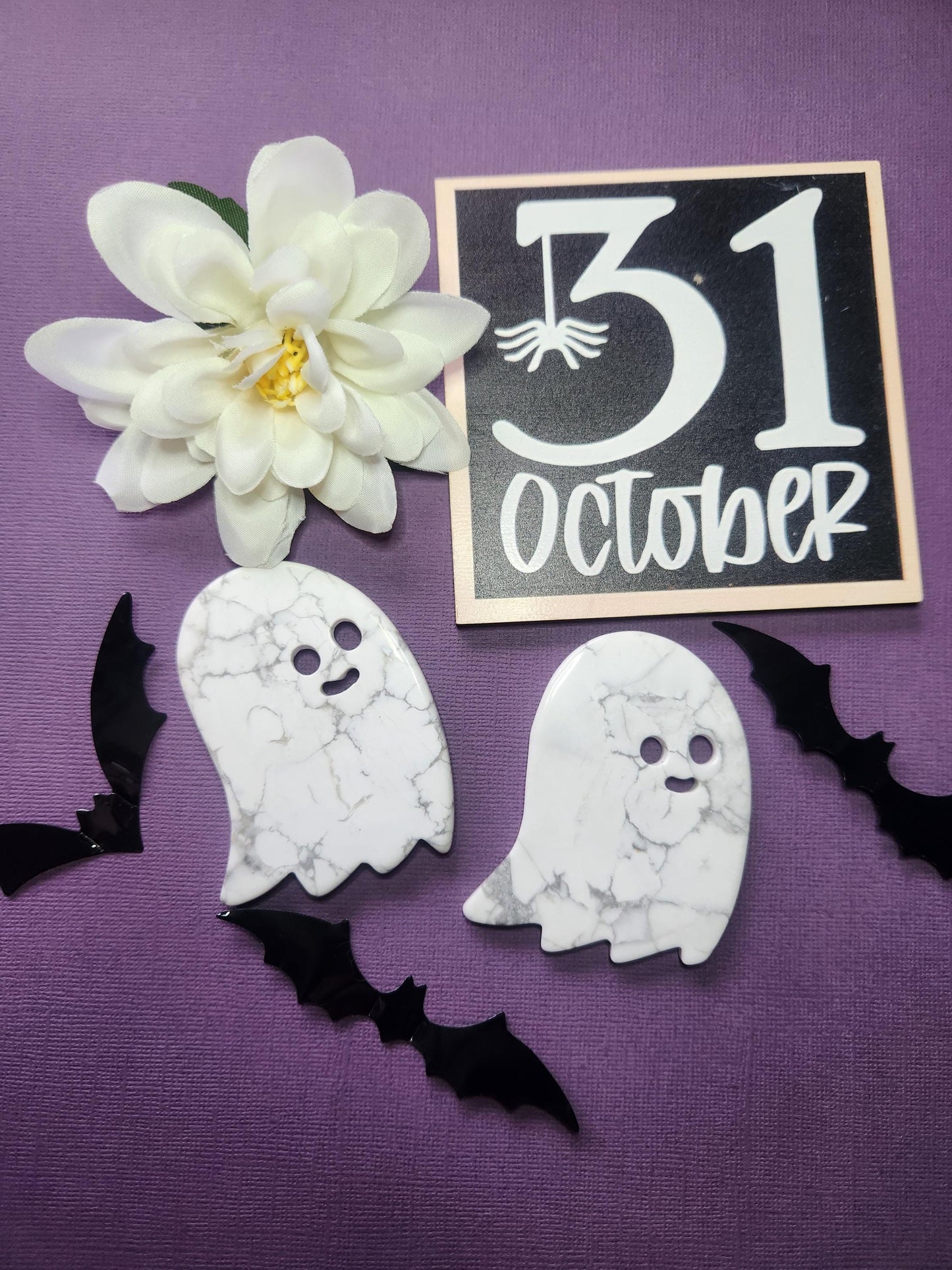 Two white howlite ghost crystal slices displayed against a dark purple backdrop, surrounded by Halloween-themed props including black bats, orange flowers, and a "31 October" sign — spooky witchy crystal decor perfect for Halloween altars and metaphysical collectors.