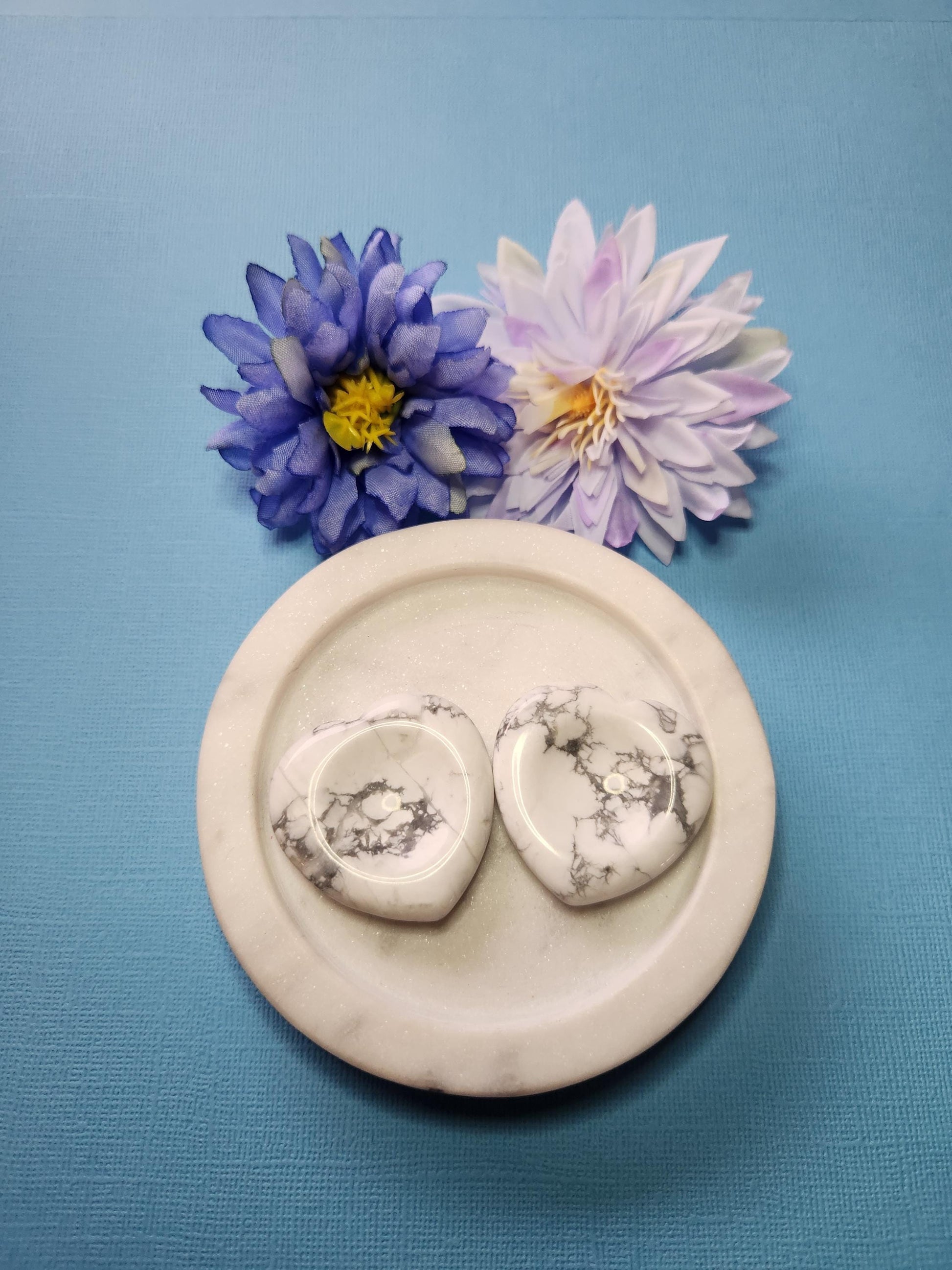 Two howlite heart-shaped worry stones displayed in a white dish alongside two flower carved crystal hearts, styled on a light blue background, calming crystal anxiety stones, soothing white howlite hearts for stress relief, aesthetic crystal shop display.