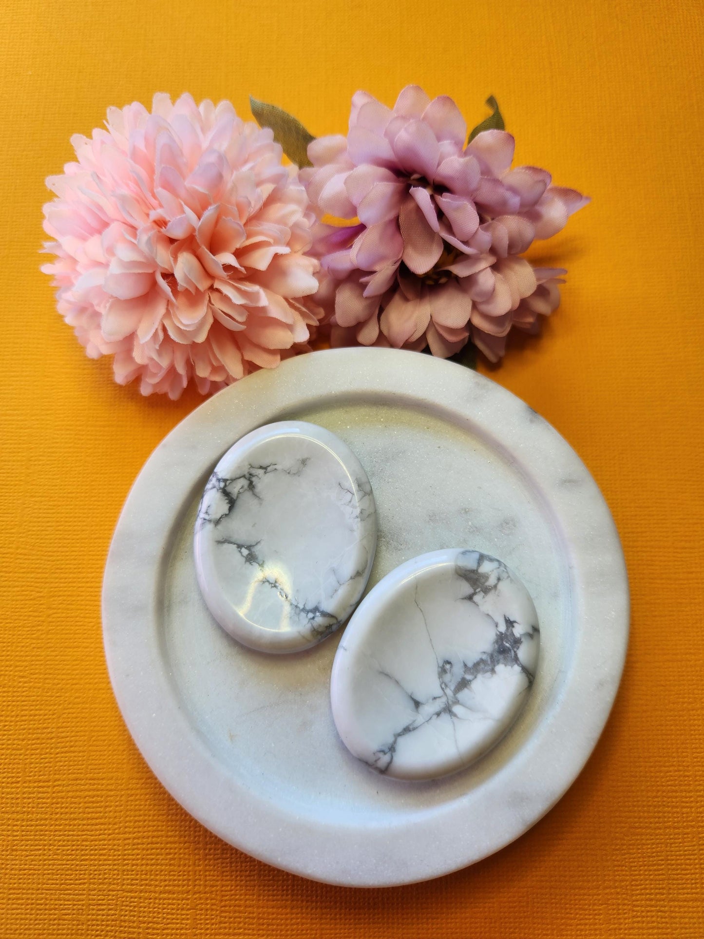 Two polished Howlite worry stones displayed on a white dish, styled with pink flower props on a bright yellow background. Natural white Howlite crystals with grey veining, perfect for stress relief, meditation, anxiety support, and calming pocket stones. Ideal for crystal collections, reiki, mindfulness, and holistic healing.