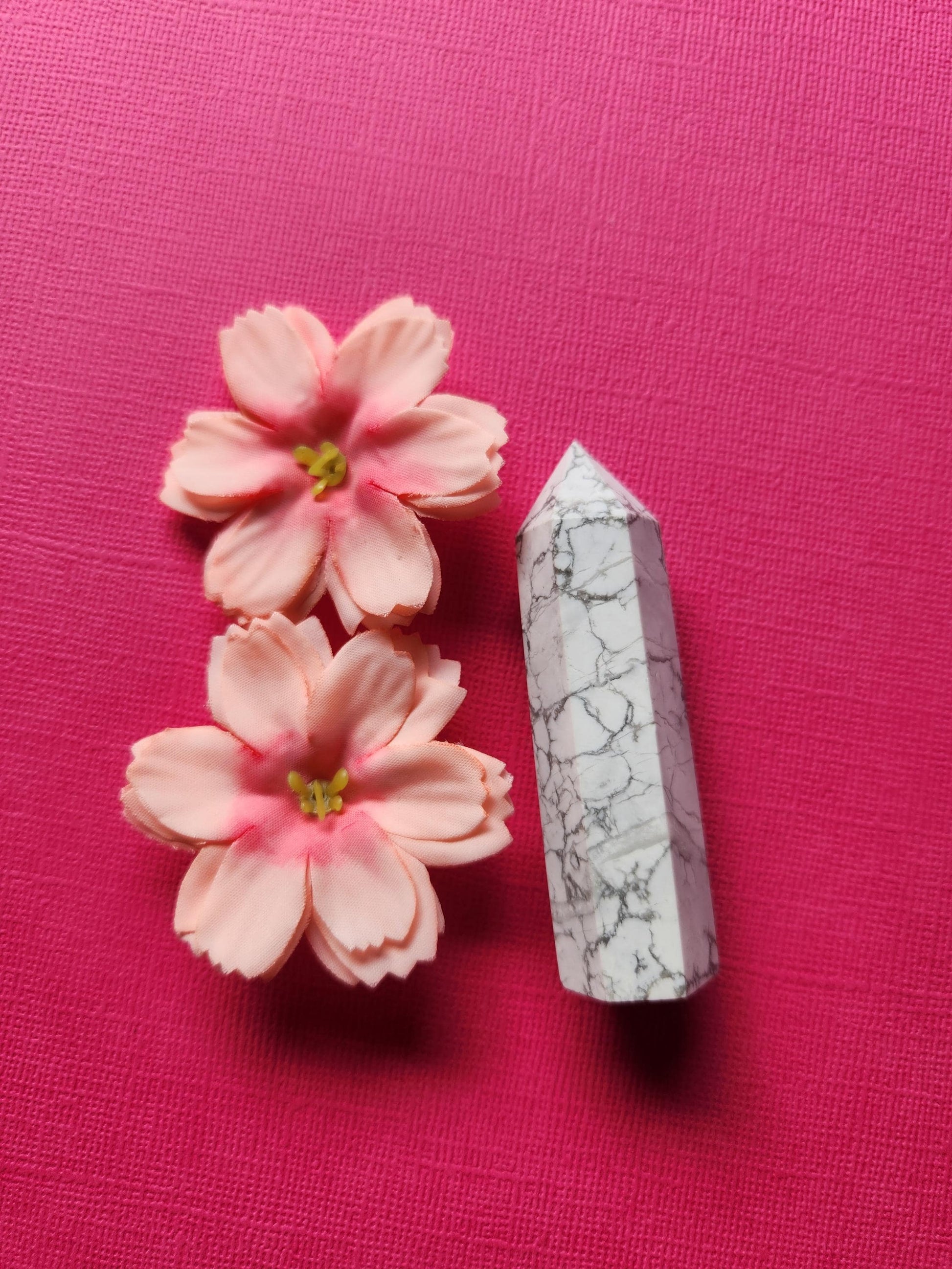 Howlite Tower standing upright against a bright dark pink background, showcasing its smooth white surface with natural gray veining. This polished crystal point is known for its calming and stress-relief properties, perfect for meditation, spiritual healing, and chakra balancing. The vibrant pink backdrop highlights the tower’s serene energy, making it an ideal addition to any crystal collection or decorative altar space.