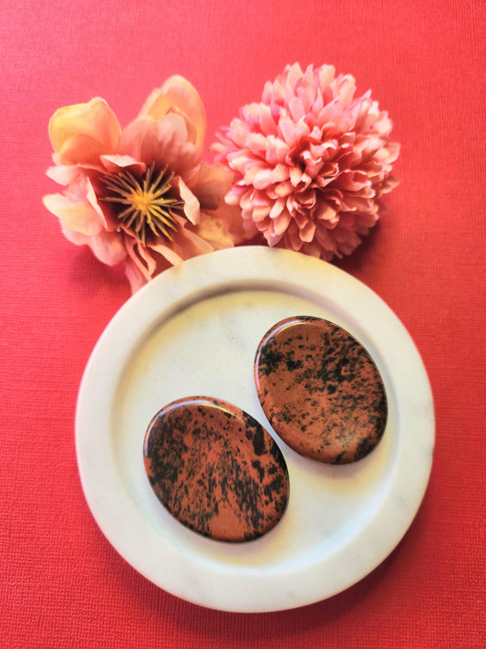 Two polished Mahogany Obsidian worry stones displayed on a white dish with two flower props, set against a red background. Deep reddish-brown and black thumb stones for grounding, protection, and strength.