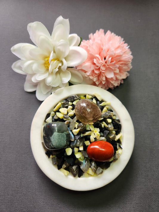 Protection Spell Confetti featuring crystal chips of Black Obsidian, Labradorite, and Serpentine, with tumbled stones of Red Jasper, Smoky Quartz, and African Bloodstone. The 50g crystal blend is displayed on a white dish with two flower props against a dark grey background, creating a grounding and powerful energy. Perfect for protection rituals, warding off negativity, energy shielding, and spiritual cleansing practices.