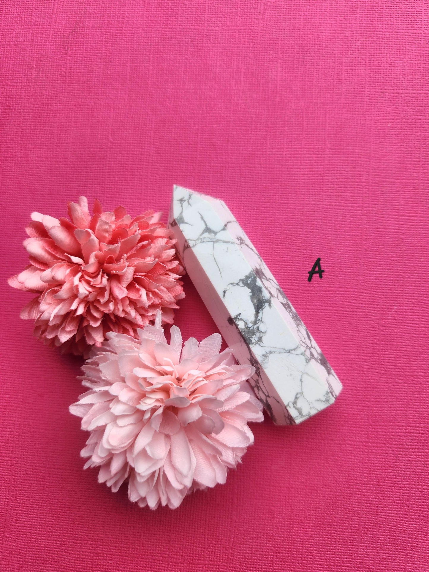 Raw white Howlite tower crystal with grey veining, polished point standing upright on a pink background with two flower props, high-quality Howlite crystal tower for anxiety relief, calming energy, meditation and crystal healing