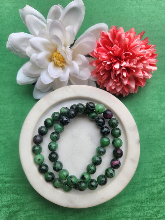 Two ruby in zoisite 8mm beaded bracelets displayed in a white dish on a green background with flower props featuring green zoisite beads with ruby inclusions for vitality growth heart chakra energy healing jewelry gift