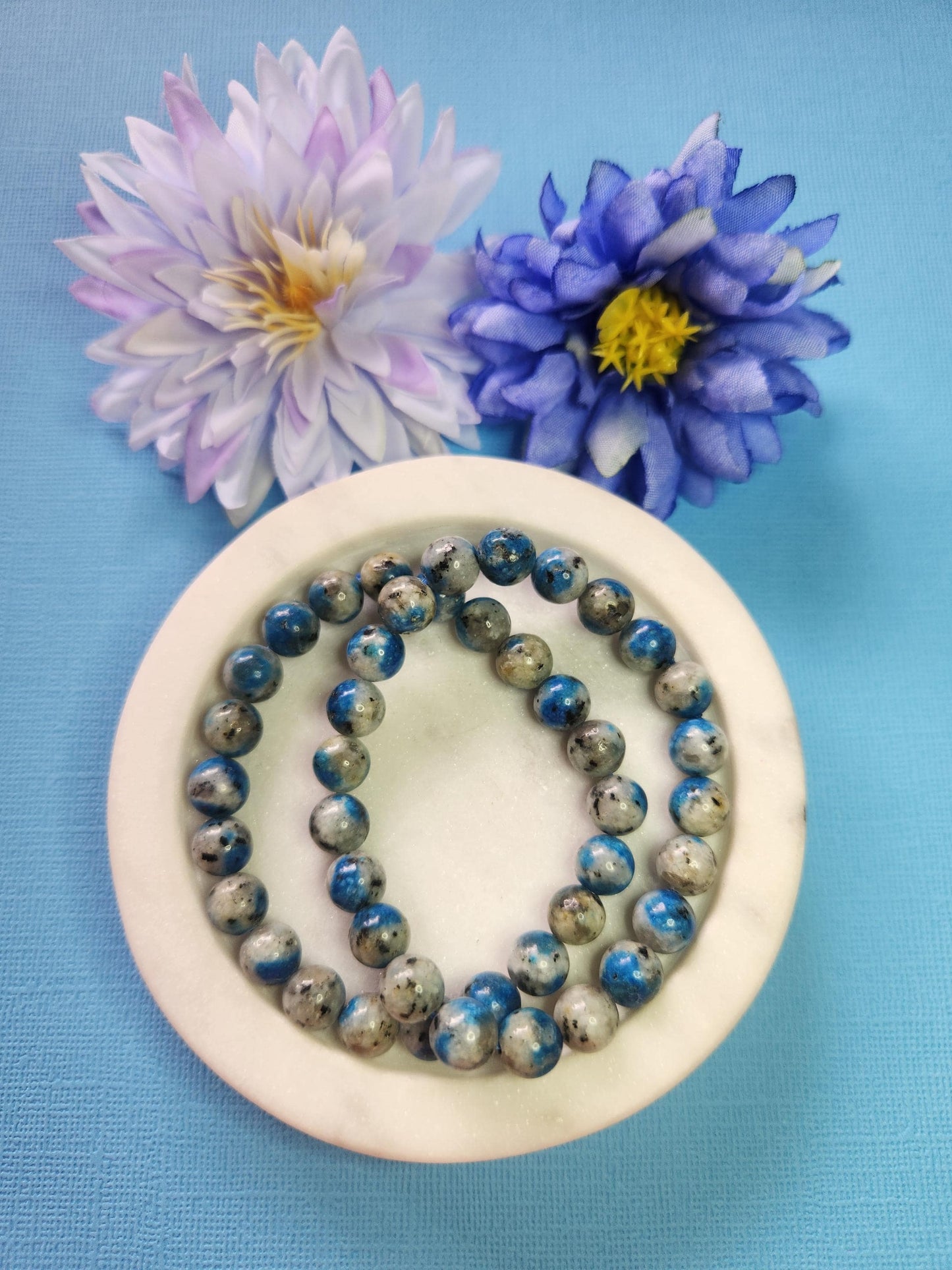 Two K2 8mm beaded bracelets in white dish with two flower props on blue background, natural K2 granite gemstone beads, handmade crystal jewellery, healing energy and grounding stone beaded bracelet product photo