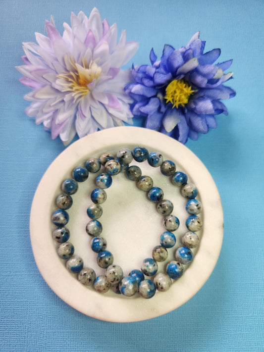 Two K2 8mm beaded bracelets in white dish with two flower props on blue background, natural K2 granite gemstone beads, handmade crystal jewellery, healing energy and grounding stone beaded bracelet product photo