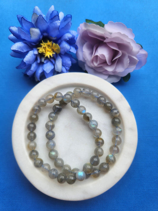 Two labradorite 8mm beaded bracelets displayed in a white dish on a blue background with soft flower props featuring polished crystal beads for protection intuition energy healing bracelet and gift