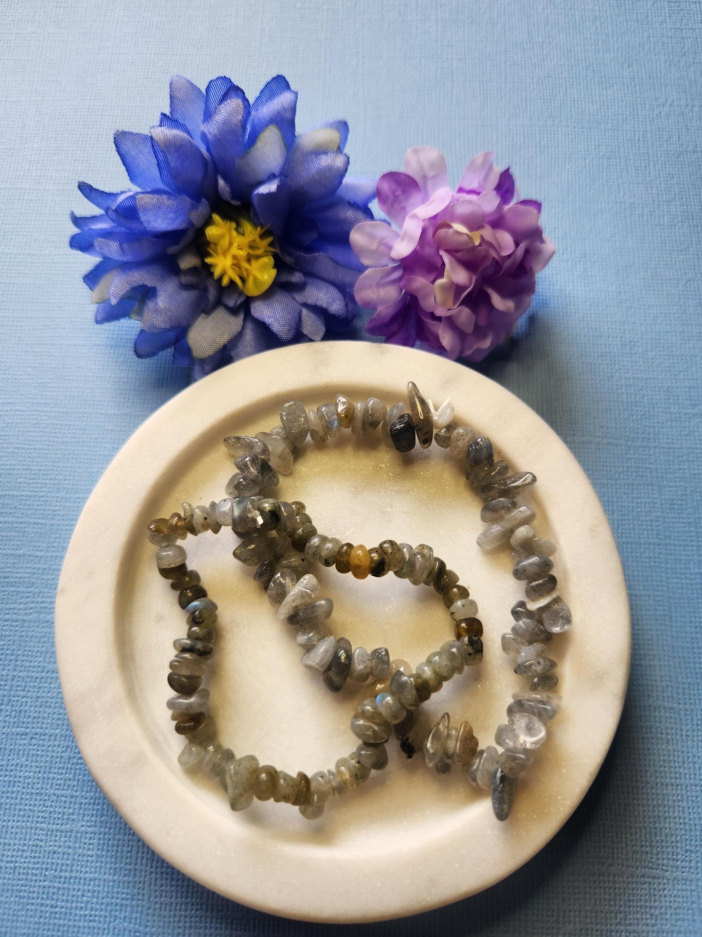 Two Labradorite chip bracelets displayed on a white dish with two blue and white flower props, set against a soft blue background. Stretch gemstone bracelets for healing, intuition, protection, chakra balancing, meditation, energy work, spiritual jewelry, mindful accessory, crystal lover gift, boho jewelry, and metaphysical energy stones