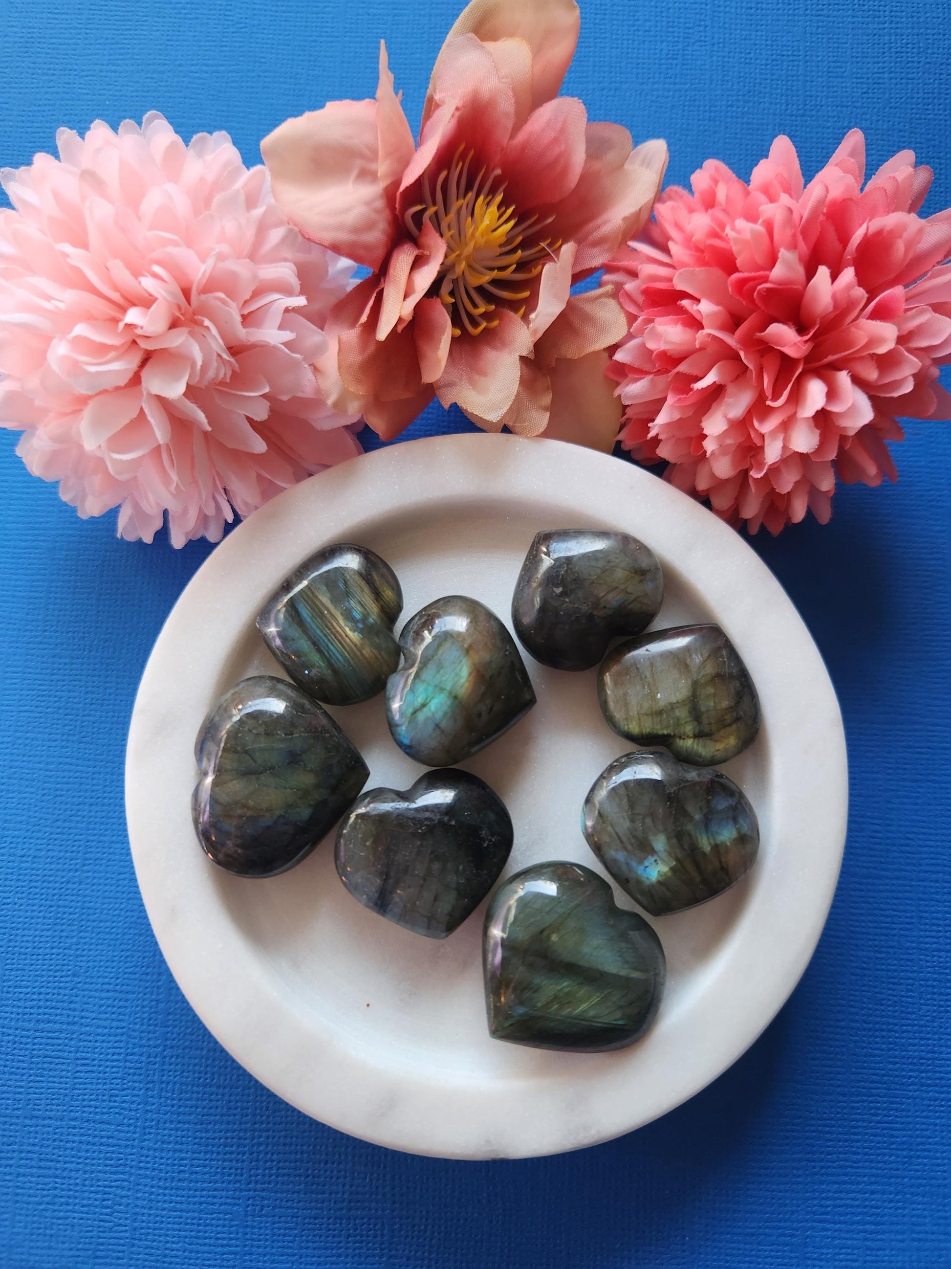Small polished Labradorite heart crystals displayed on a blue background. These natural gemstone hearts feature beautiful flashes of blue, green, and gold, known for their protective and intuitive energy. Ideal for chakra balancing, meditation, spiritual healing, and as pocket stones. Labradorite supports transformation and shielding negative energy. Perfect for gifting, energy work, and crystal collectors.