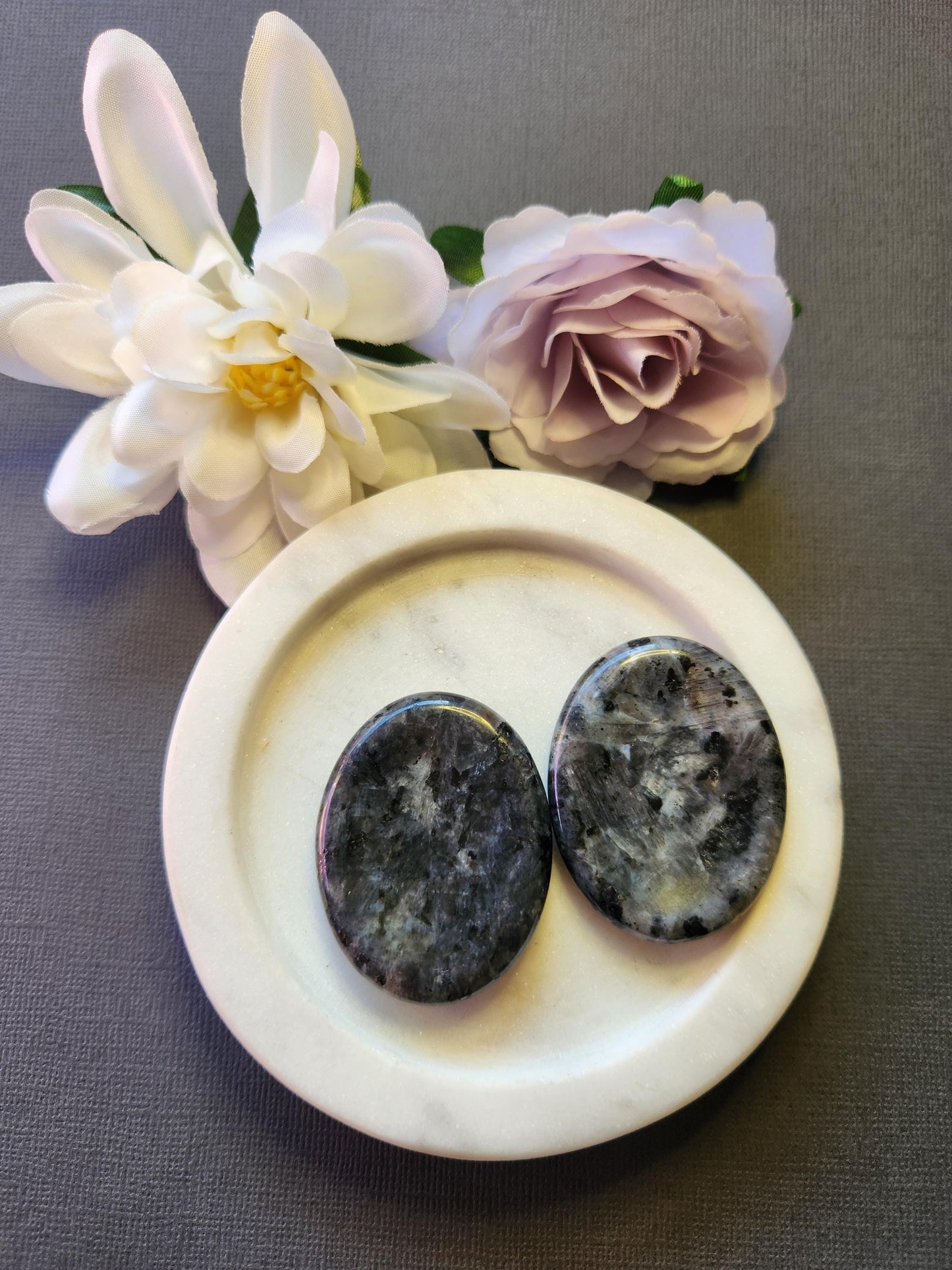 Two polished Larvikite worry stones displayed on a white dish with two flower props, set against a grey background. Dark grey thumb stones with subtle flashes, used for grounding, protection, and cleansing energy.