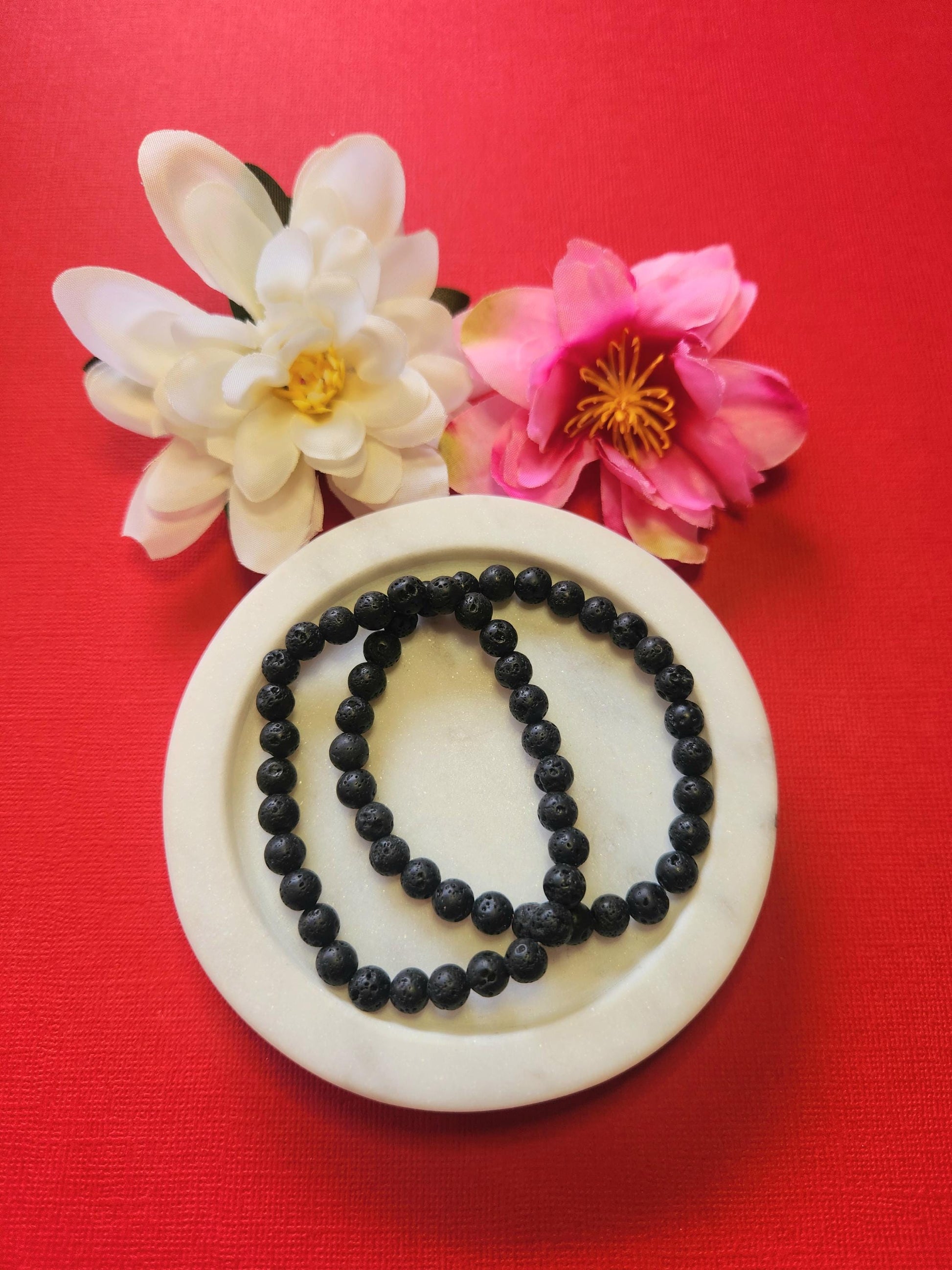 Photo showcasing two Lava Stone 6mm beaded bracelets displayed in a white dish with two flower props on a red background, highlighting the polished black crystal beads, grounding energy, and diffuser-friendly properties — perfect for aromatherapy, meditation, and crystal jewelry stacking.
