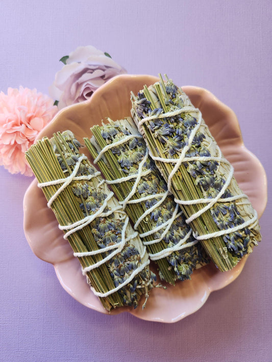 Three sage and lavender smudge sticks displayed in a pink flower-shaped bowl with two flower props, set against a lavender background. Hand-tied natural herbs for cleansing, energy clearing, relaxation, and creating a harmonious, peaceful environment — perfect for spiritual practices and gifting.