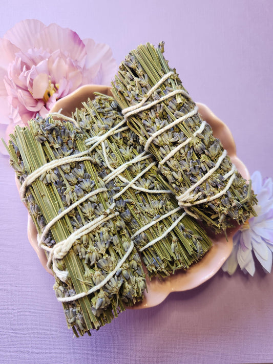 Three lavender smudge sticks displayed in a pink flower-shaped bowl with two flower props, set against a lavender background. Hand-tied natural lavender for calming, relaxation, energy clearing, and spiritual practices — perfect for meditation, home rituals, and gifting.