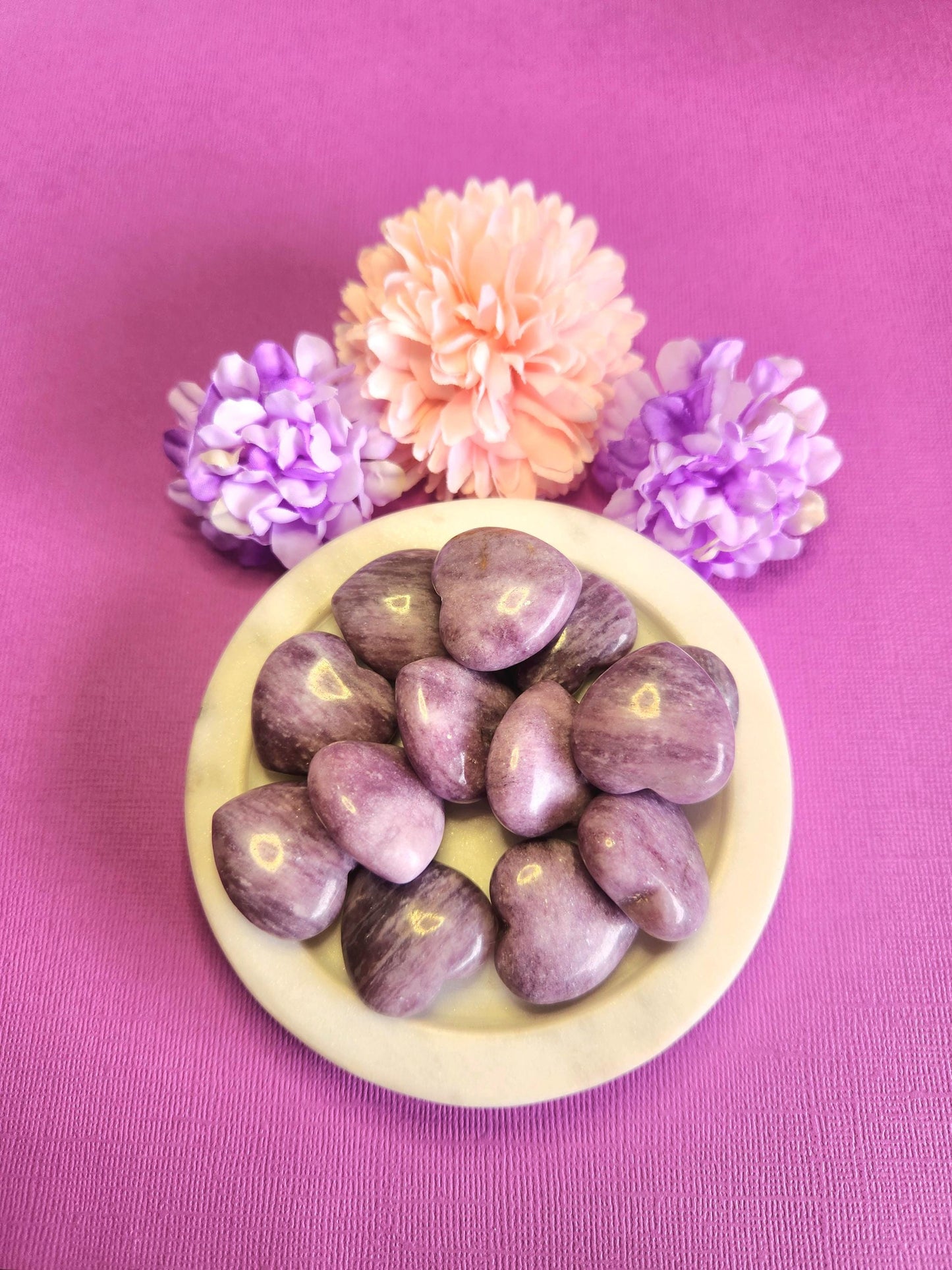 Lepidolite crystal hearts displayed in a white dish on a purple background with soft flower props featuring polished purple heart stones for calming stress relief emotional balance spiritual healing crystal decor and gift