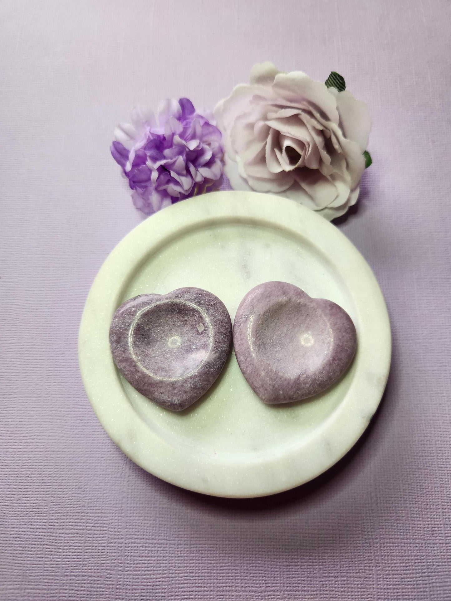Two Lepidolite heart worry stones in a white dish with two flowers on a lavender background, polished semi-precious crystals for anxiety relief, meditation, and spiritual healing.