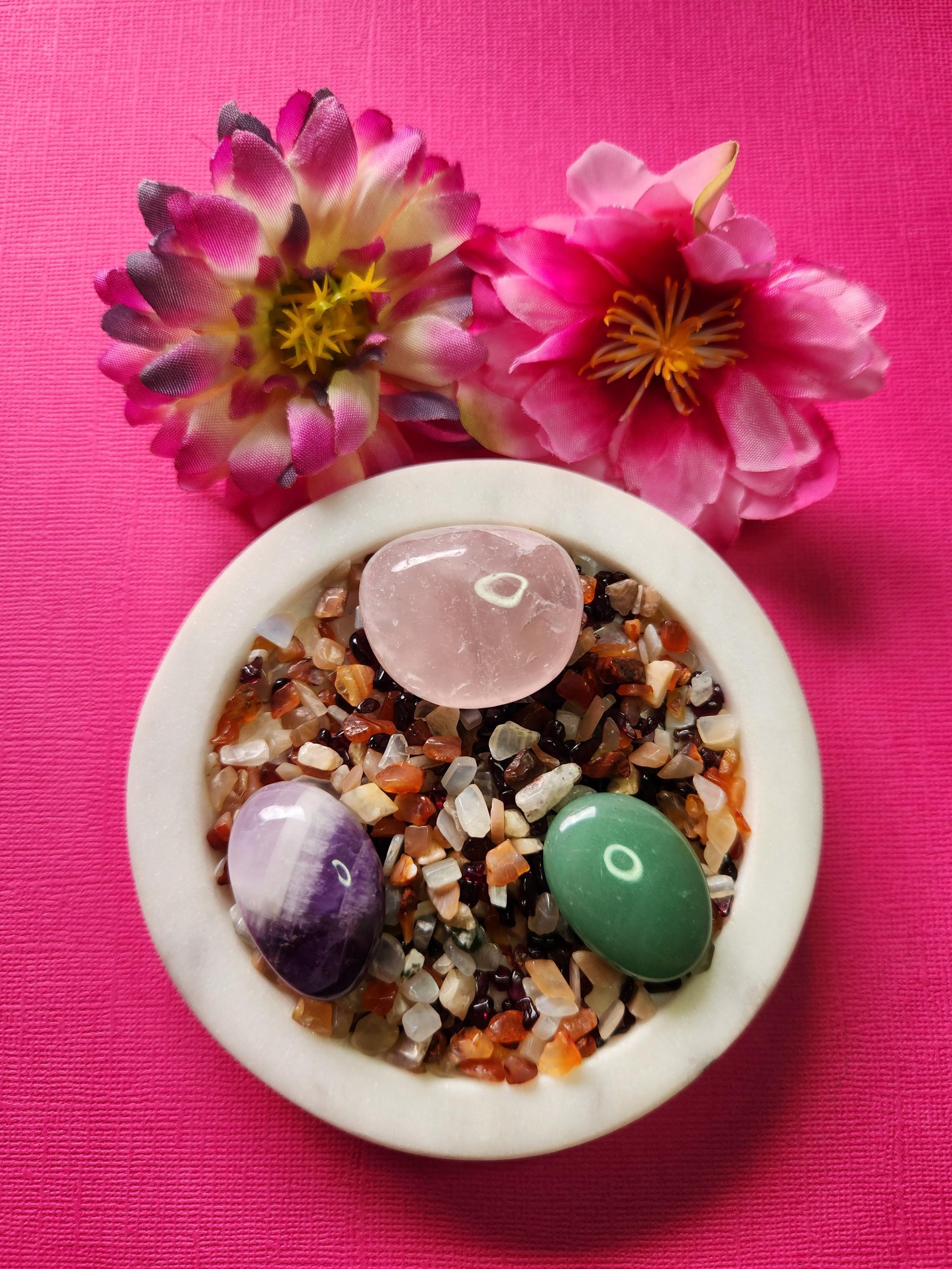 Love Spell Confetti featuring crystal chips of Carnelian, Peach Moonstone, and Garnet, with tumbled stones of Rose Quartz, Green Aventurine, and Amethyst. The vibrant crystal mix is displayed on a white dish with two flower props against a soft pink background, radiating warm, heart-opening, and romantic energy. Perfect for self-love rituals, attraction spells, emotional healing, and crystal manifestation practices.