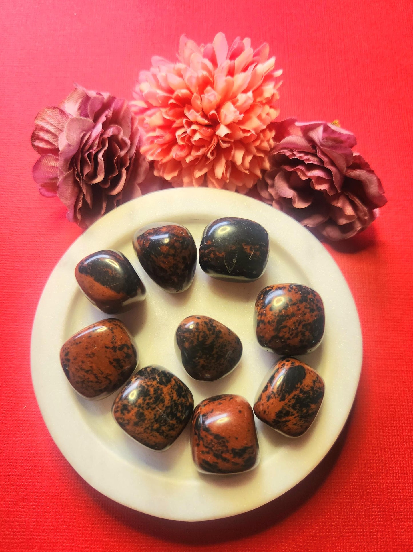 Mahogany obsidian tumble stones displayed in a white dish with three flower props, set against a red background. Polished natural obsidian pieces known for grounding, strength, and protection — perfect for crystal collections, meditation, and spiritual décor.