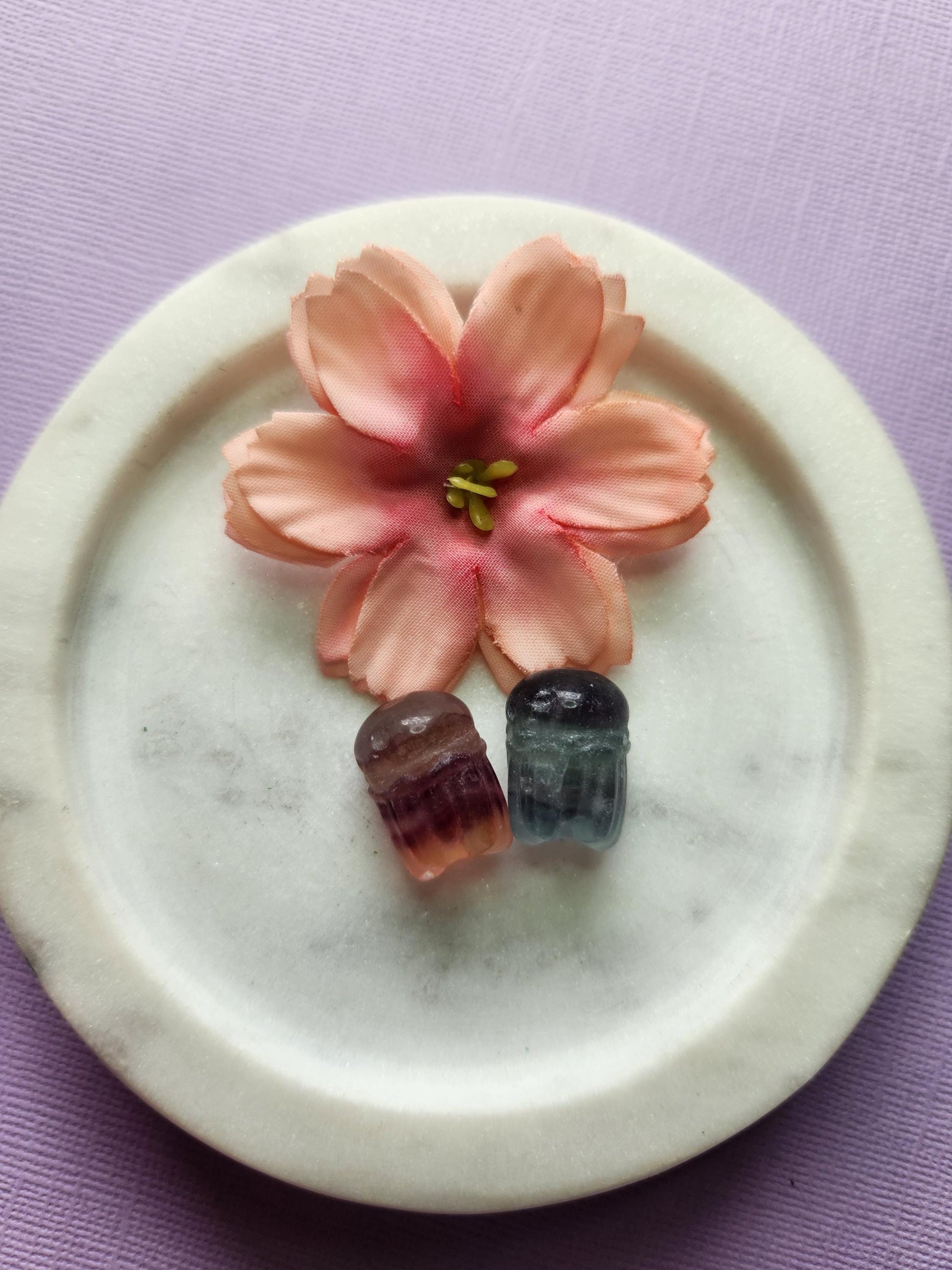 two fluorite mini octopus carvings sitting in a white ceramic dish, surrounded by two soft floral props on a lavender backdrop. The mini octopus carvings show swirling bands of purple, green and blue fluorite, polished with a glossy shine. Perfect for crystal collectors, fluorite carvings, octopus crystal decor and cute mini crystal accents