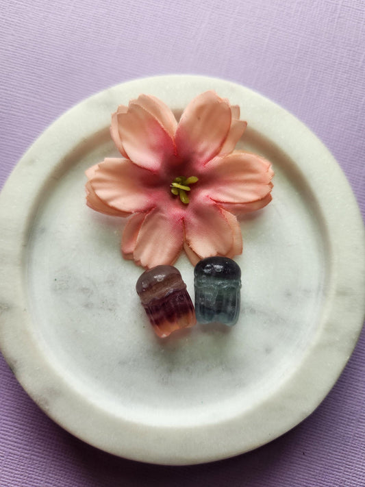two fluorite mini octopus carvings sitting in a white ceramic dish, surrounded by two soft floral props on a lavender backdrop. The mini octopus carvings show swirling bands of purple, green and blue fluorite, polished with a glossy shine. Perfect for crystal collectors, fluorite carvings, octopus crystal decor and cute mini crystal accents