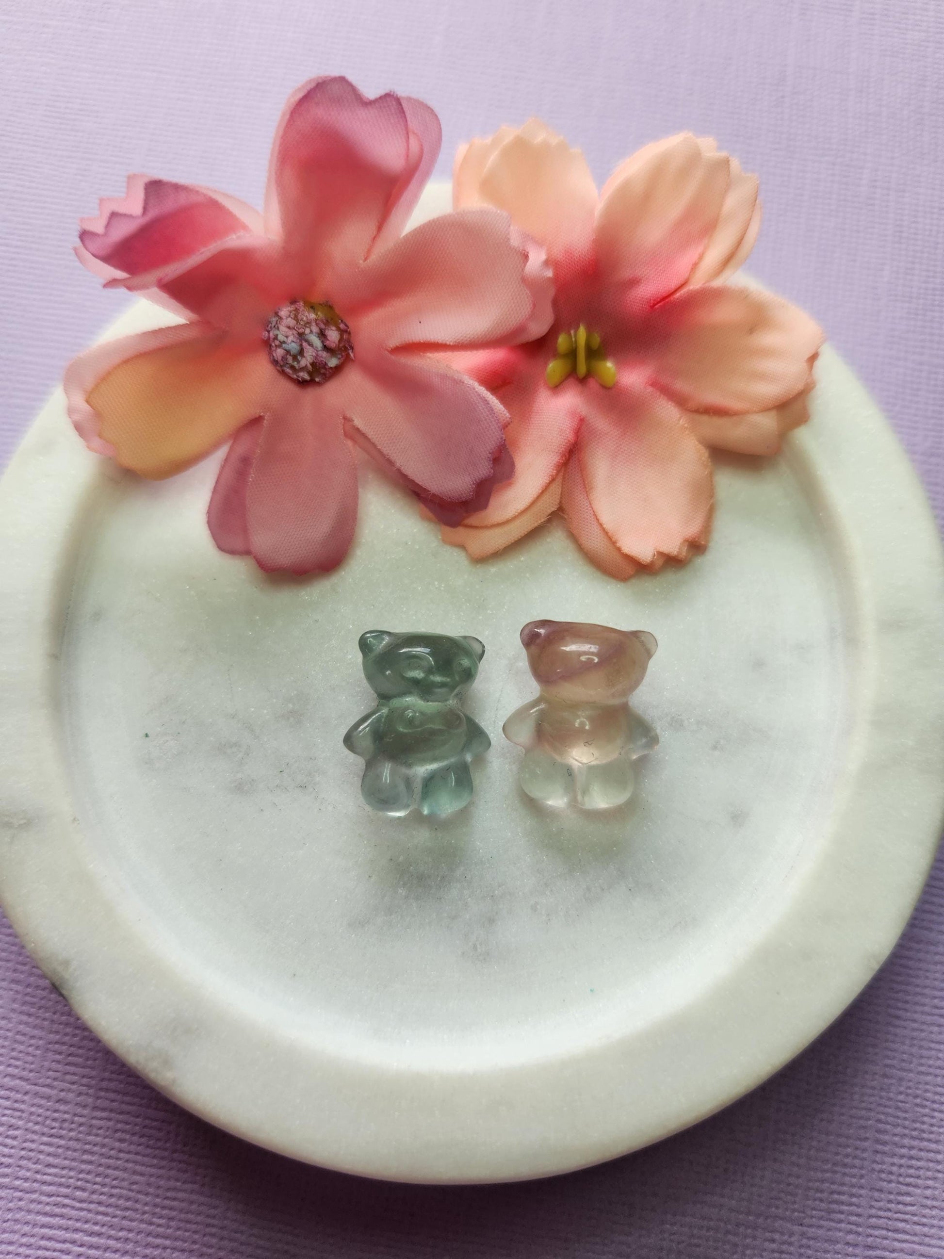 Close-up photo of two fluorite mini bear carvings displayed in a white dish with two soft flower props on a lavender backdrop. The bears show bands of purple, green and blue fluorite with a polished shine. Ideal for fluorite carvings, mini crystal animals, bear crystal decor and cute collectible gemstones