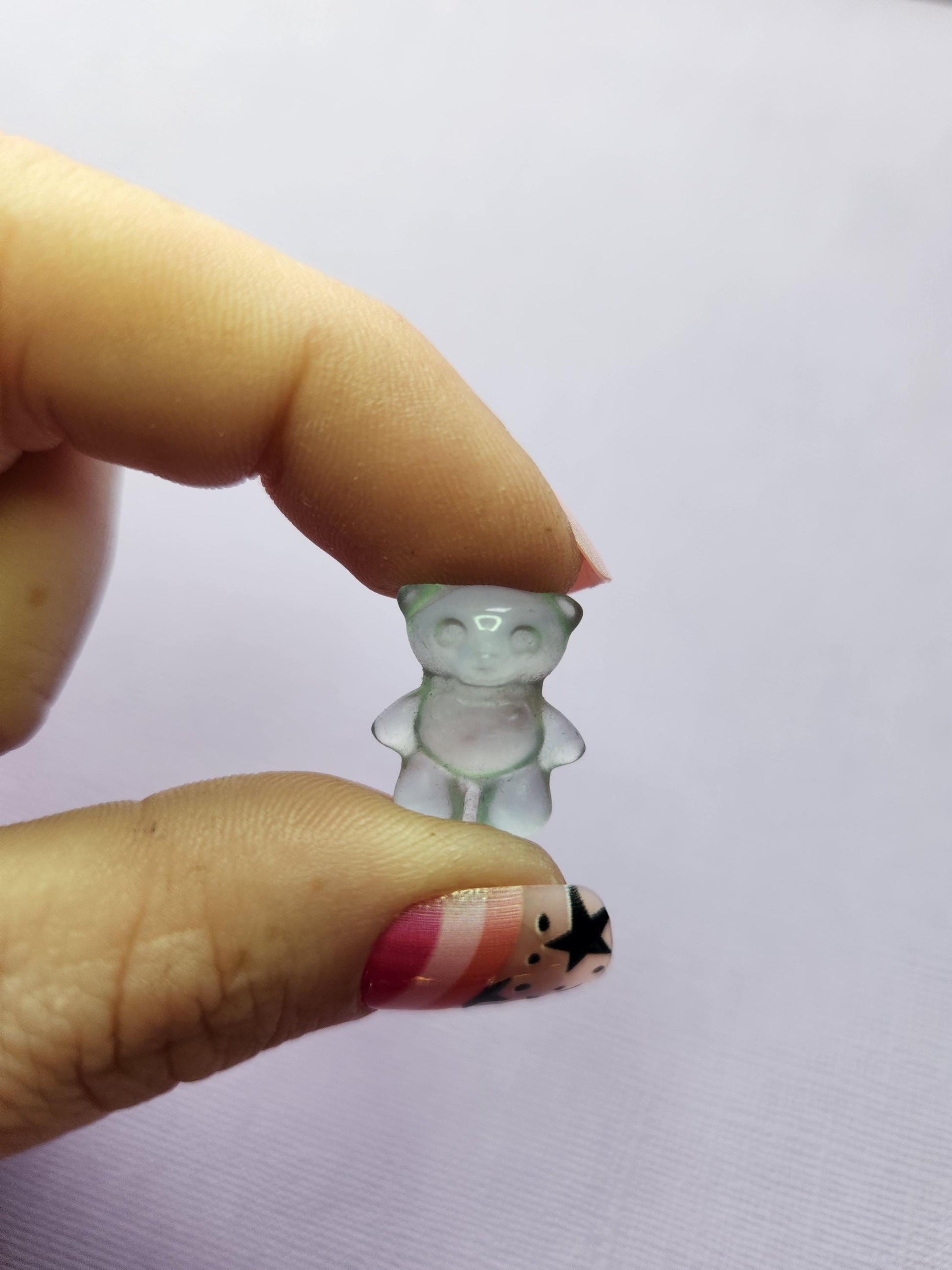 Close-up photo of a single fluorite mini bear carving held between fingers, showing detailed shaping and natural bands of purple, green and blue fluorite. Focused against a lavender backdrop. Perfect for fluorite carvings, mini crystal animals, bear crystal collectors and pocket-sized gemstone companions