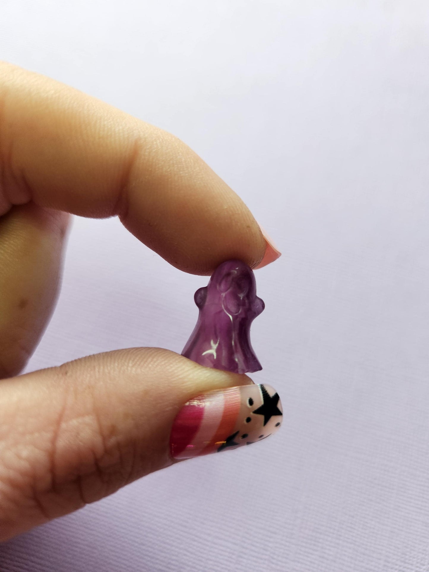 Close-up photo of a single fluorite mini ghost carving held between fingers, showing smooth polished details and natural purple, green and blue fluorite banding against a lavender backdrop. Perfect for fluorite carvings, mini crystal ghosts, collectible gemstone figures and pocket-sized crystals